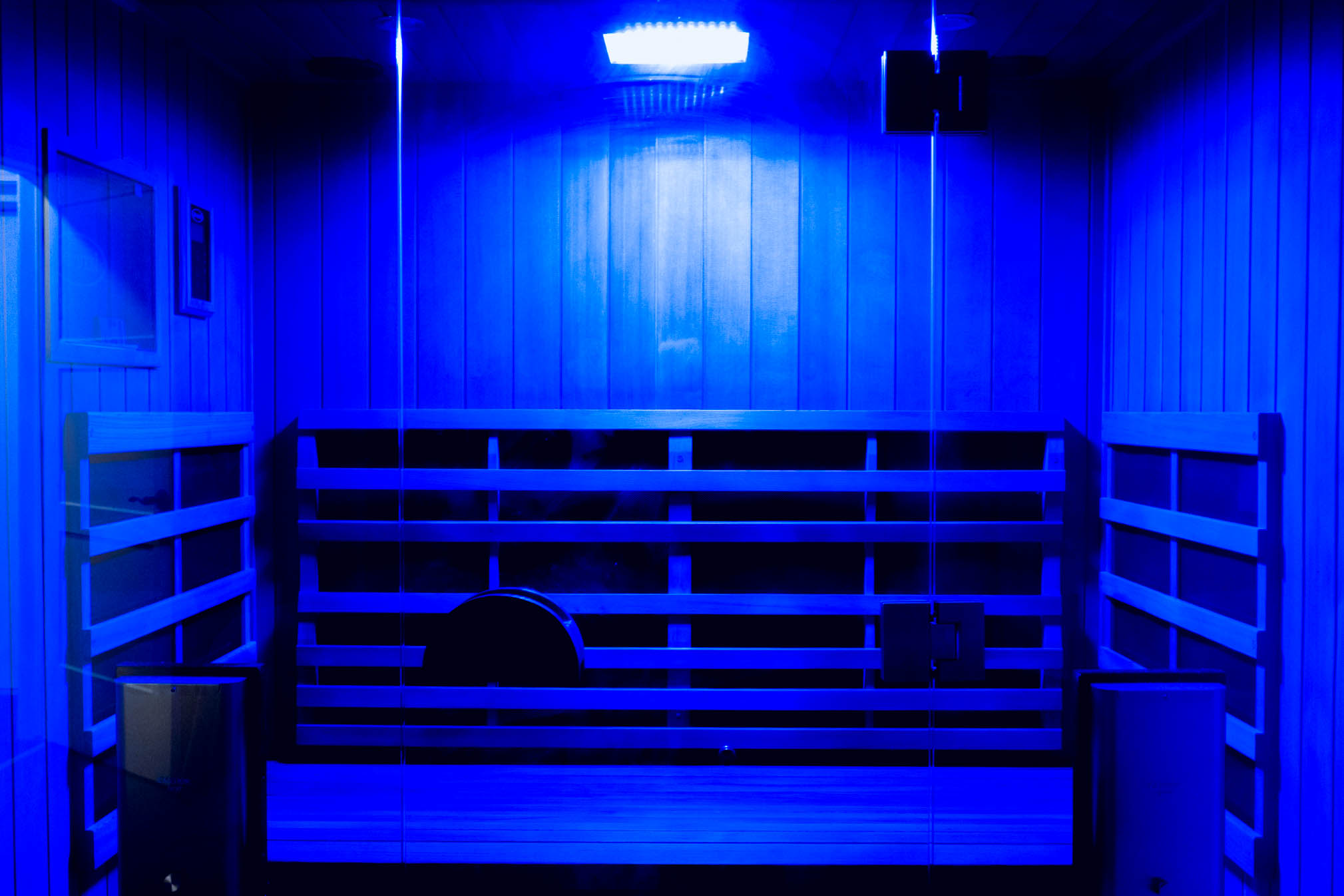 Empty wooden bench inside a small room illuminated by blue light, with wooden walls and a ceiling light.
