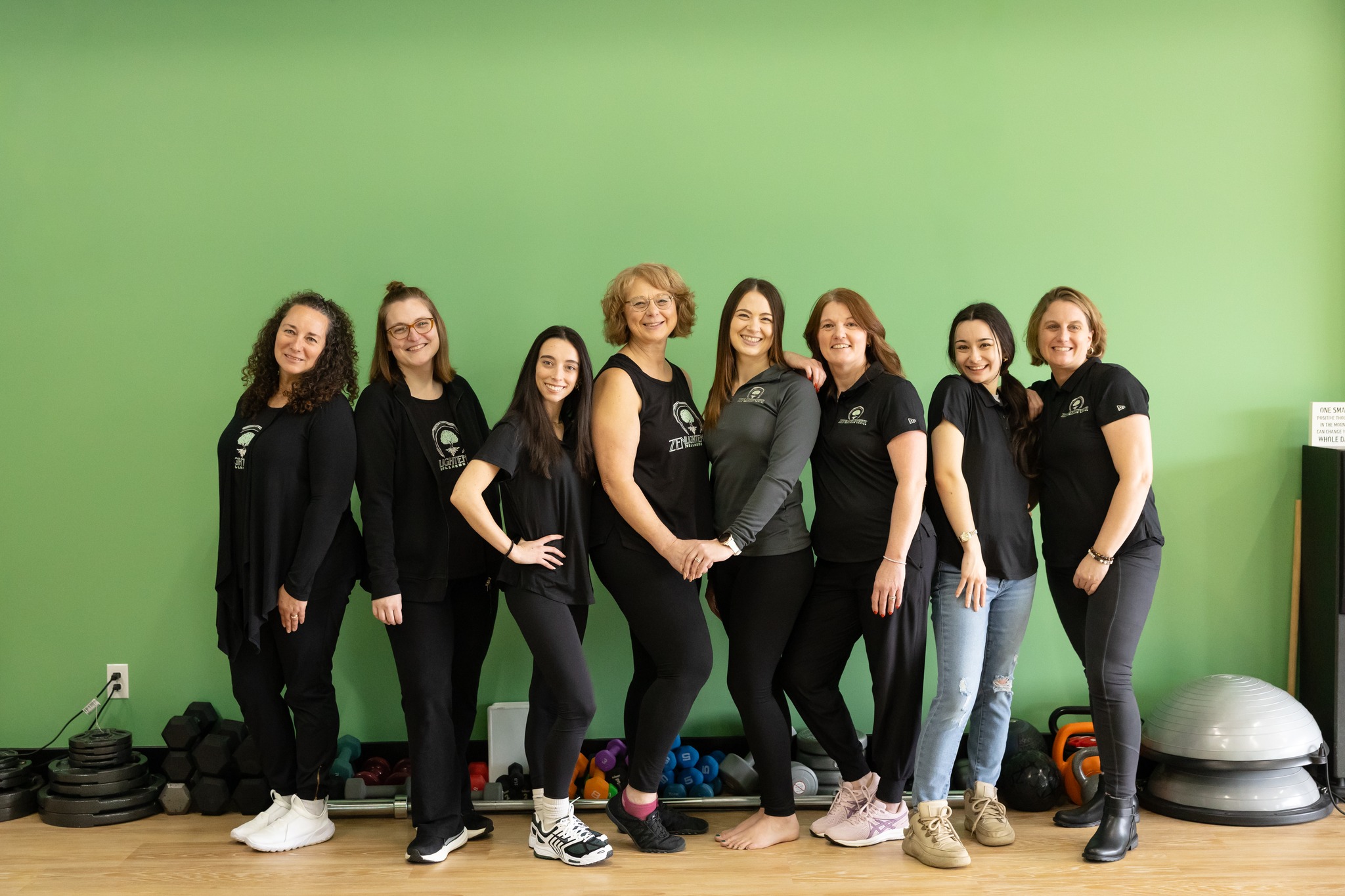 Group of eight women standing together in front of a green wall, smiling, wearing black shirts and pants.