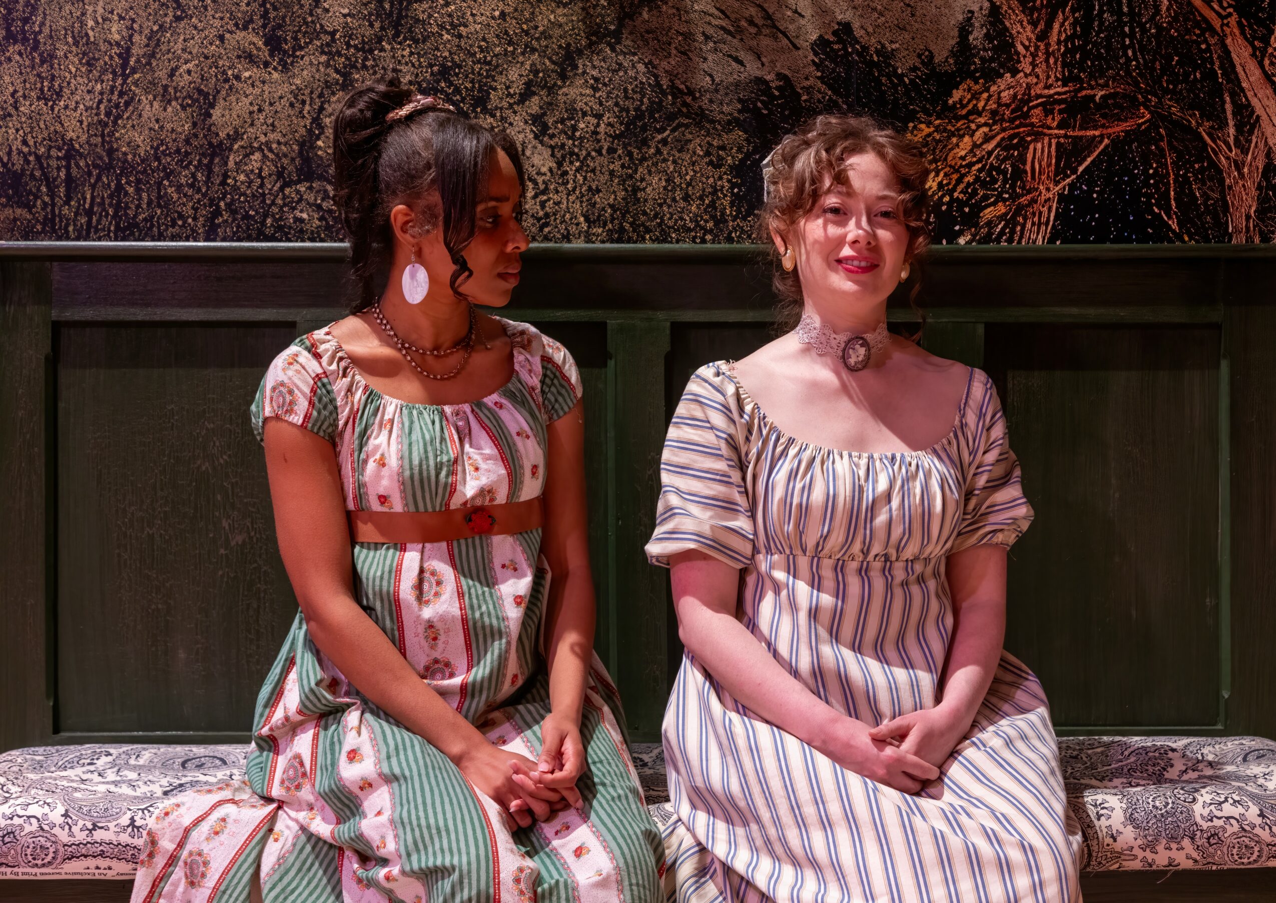 Two women sitting on a bench, one with dark skin and long hair, the other with light skin and short hair, smiling.