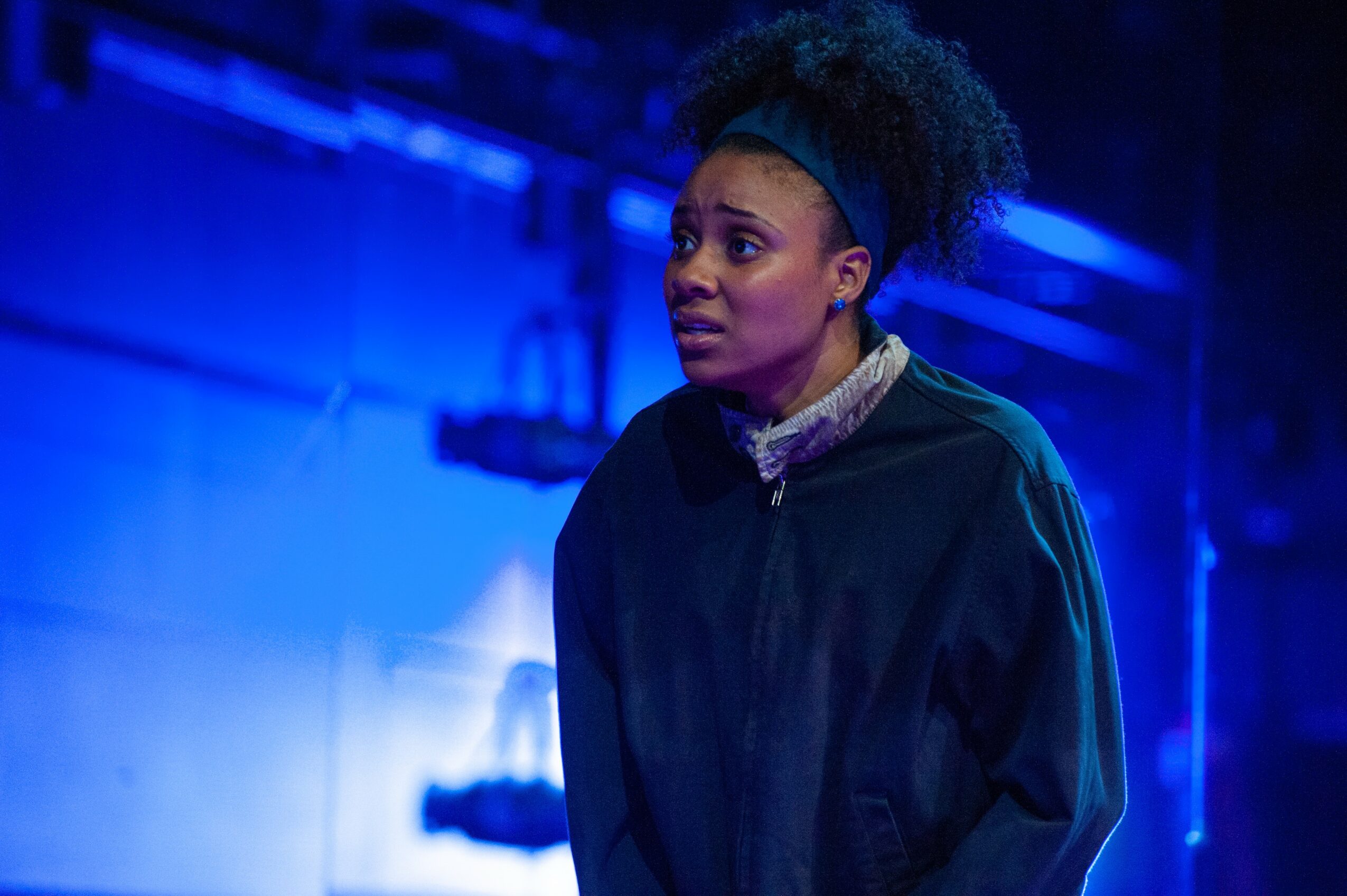 Young woman with curly hair tied back, wearing a dark jacket and headband, standing in a blue-lit environment.