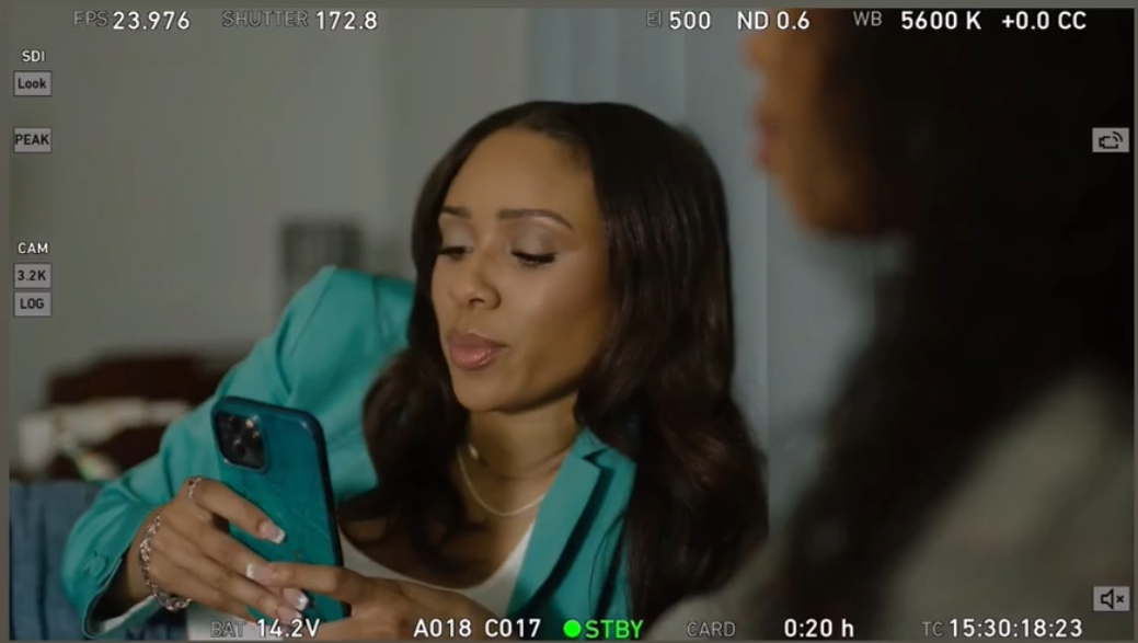 Woman with long dark hair looking at her phone, wearing a teal jacket, in a room with a blurred person nearby.