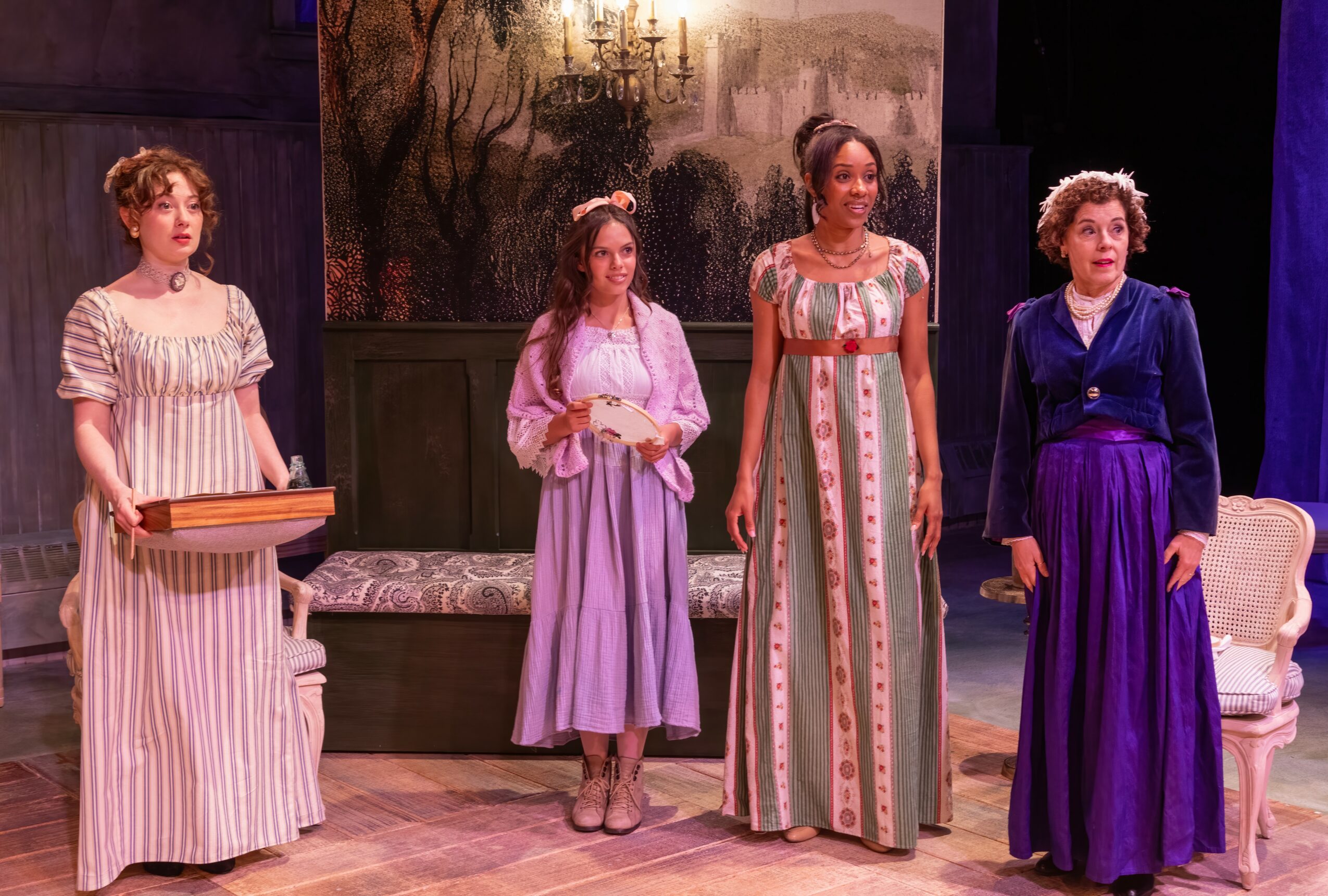 Four women in period costumes on stage, with a painted backdrop and furniture, performing in a play.