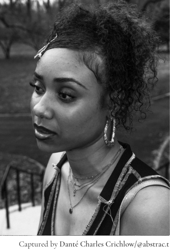 Woman with curly hair, hoop earrings, layered necklaces, and sleeveless top outdoors, looking to the side.