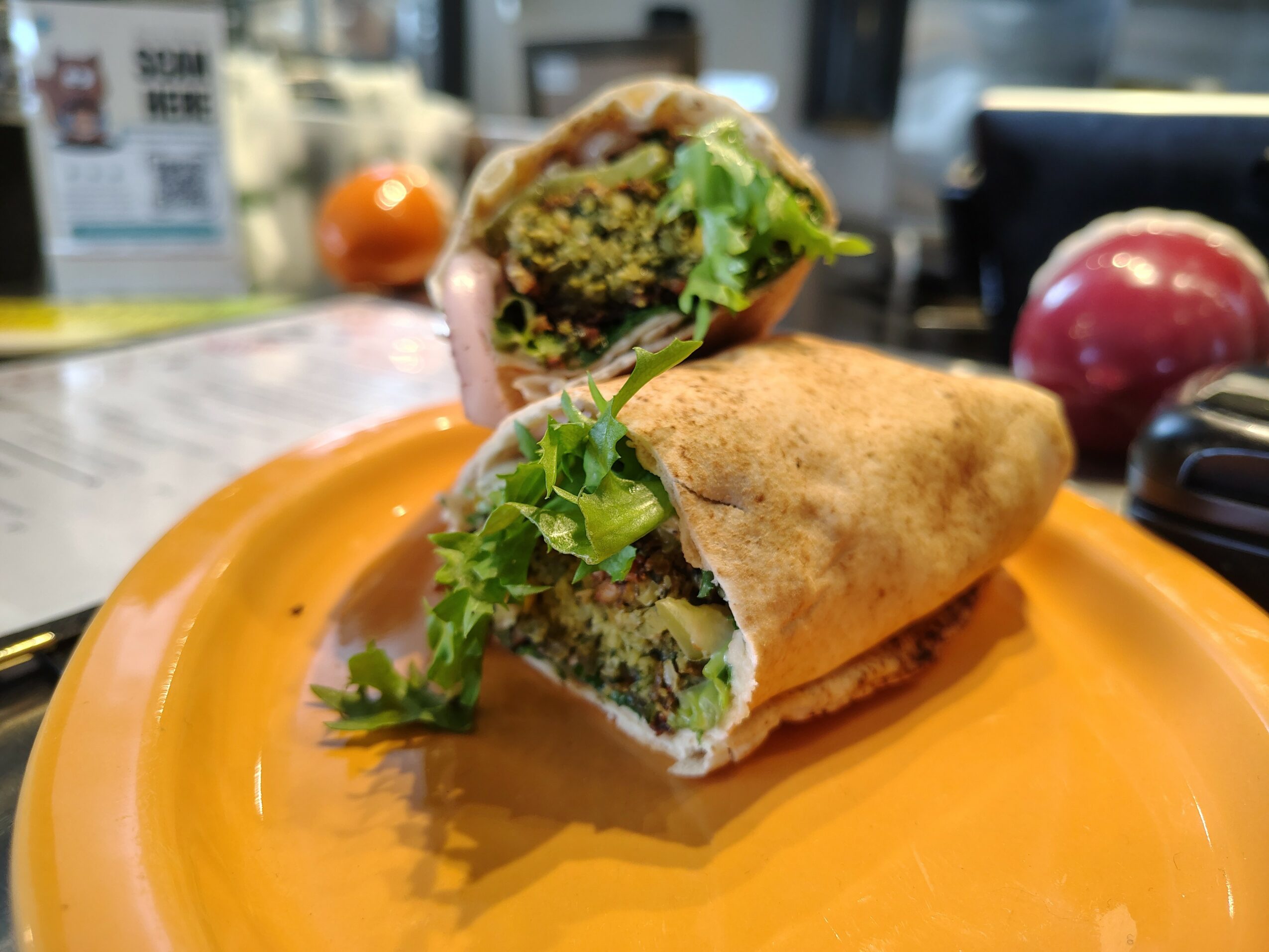 Half-eaten wrap with green filling and leafy greens on an orange plate, with a blurred background including a red onion.