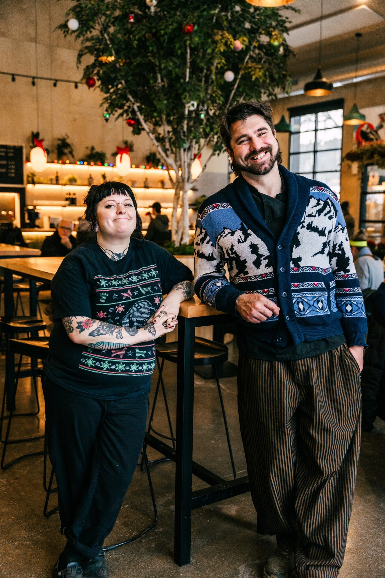 Two people standing in a cafe with a large tree and shelves in the background, smiling at the camera.