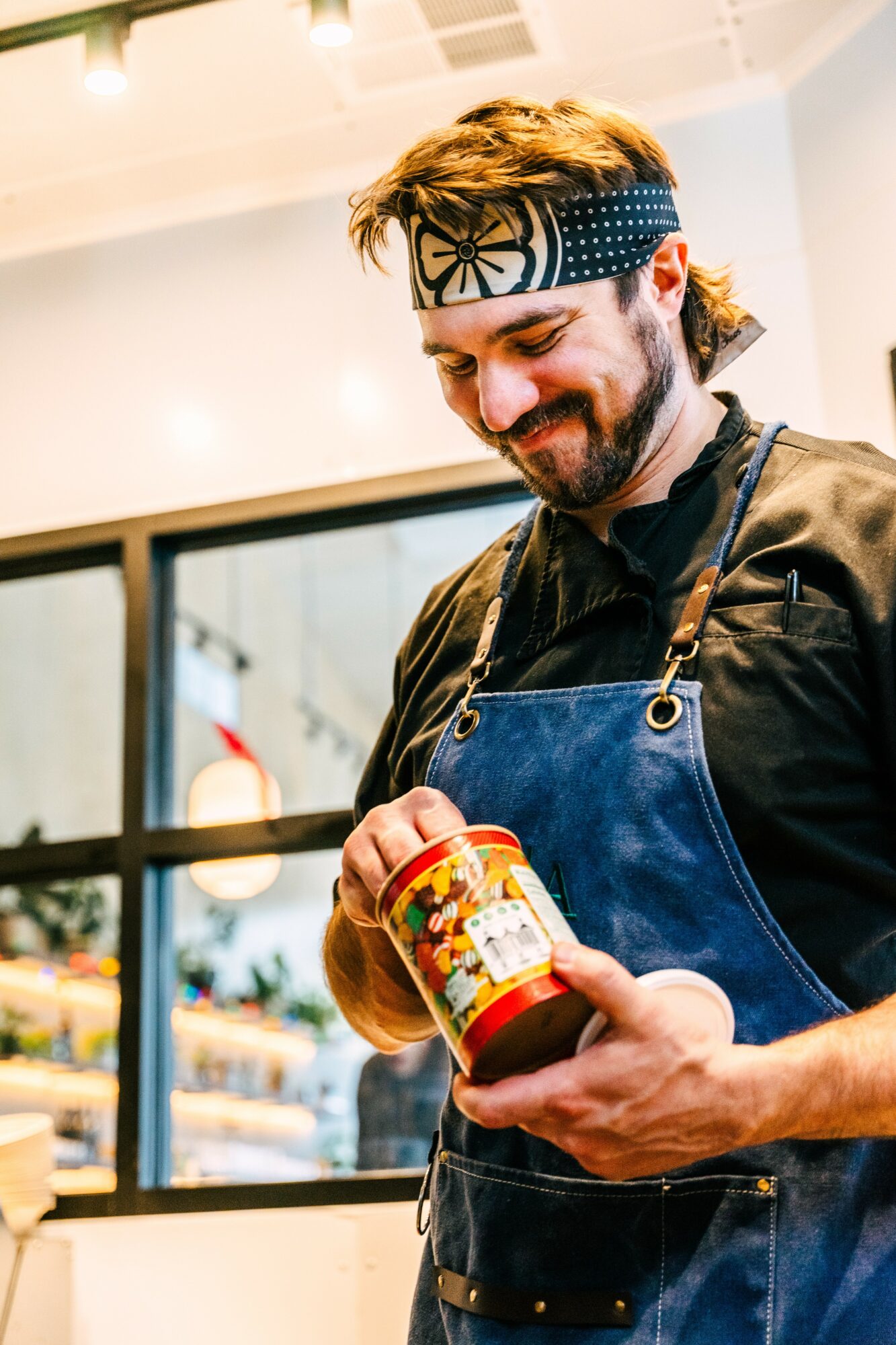Man with a headband and apron holding a jar, smiling, in a bright indoor setting.