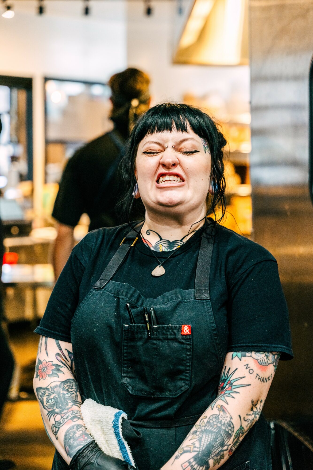 Woman with black hair and tattoos making a grimace, wearing a black apron inside a cafe or restaurant.
