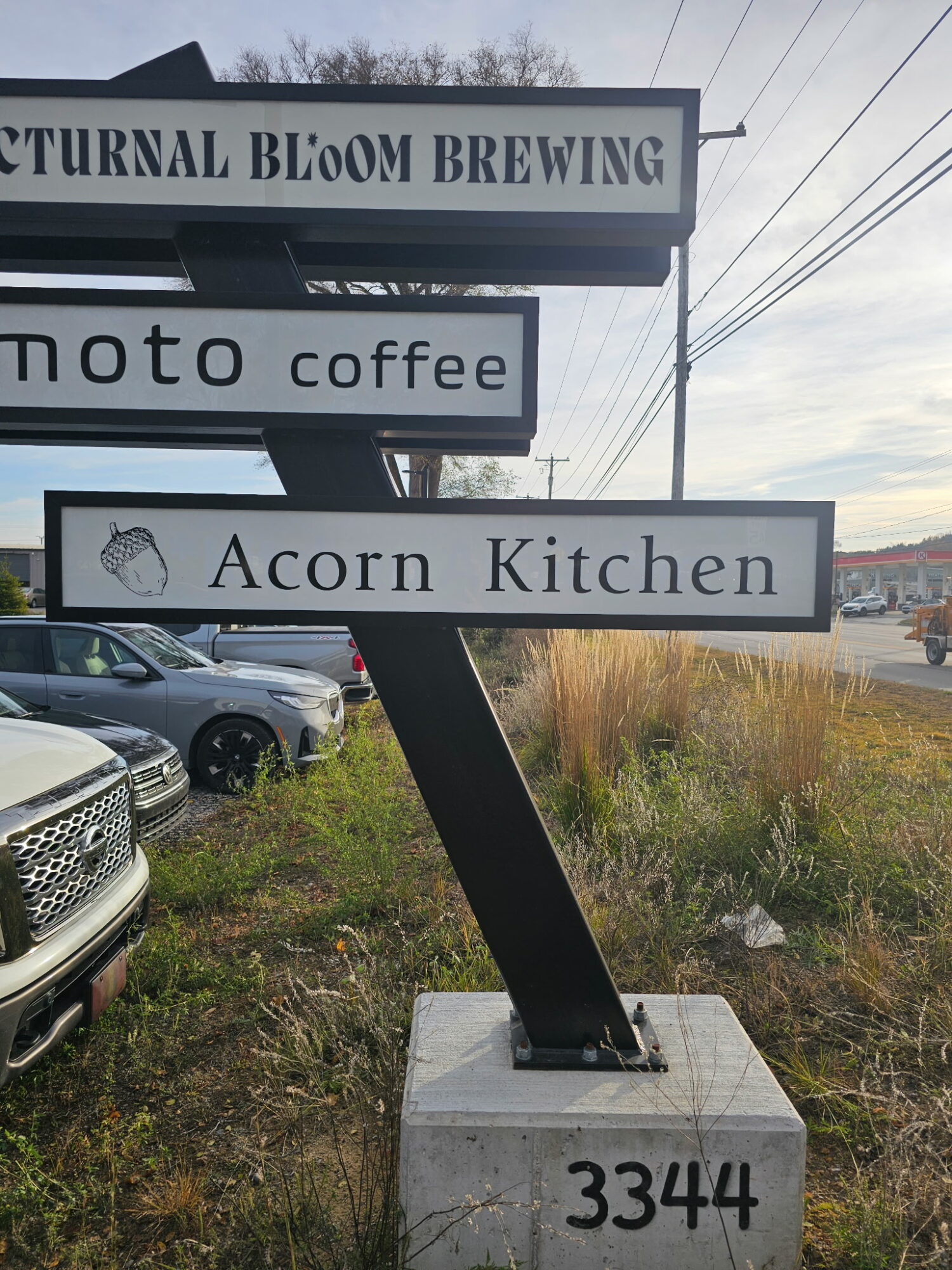 Signpost with three signs reading 'TURNTAL BLOOM BREWING', 'moto coffee', and 'Acorn Kitchen', near parked cars and grassy area.
