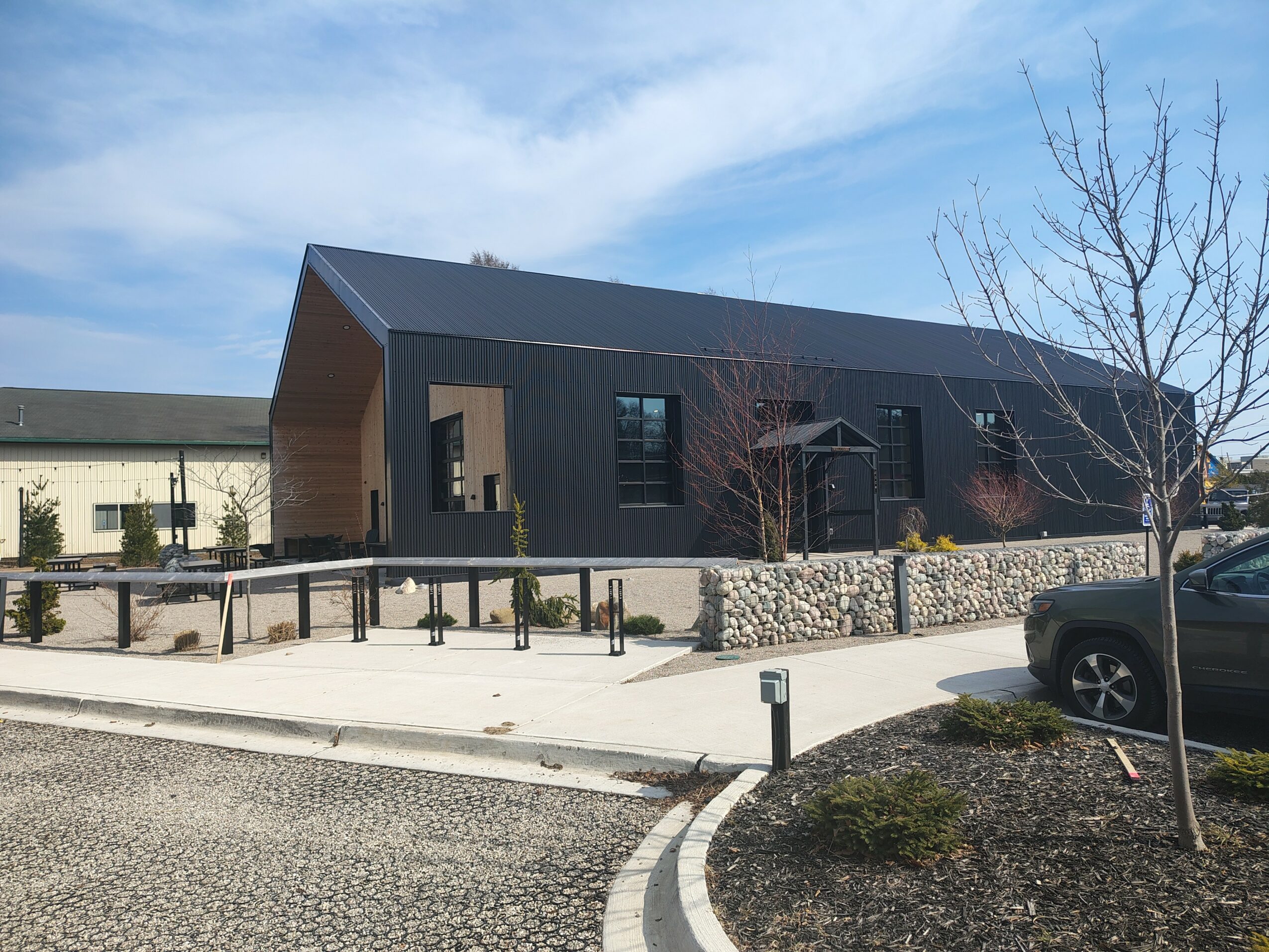 Modern building with dark exterior, large windows, and a sloped roof, surrounded by a stone wall and a paved walkway.