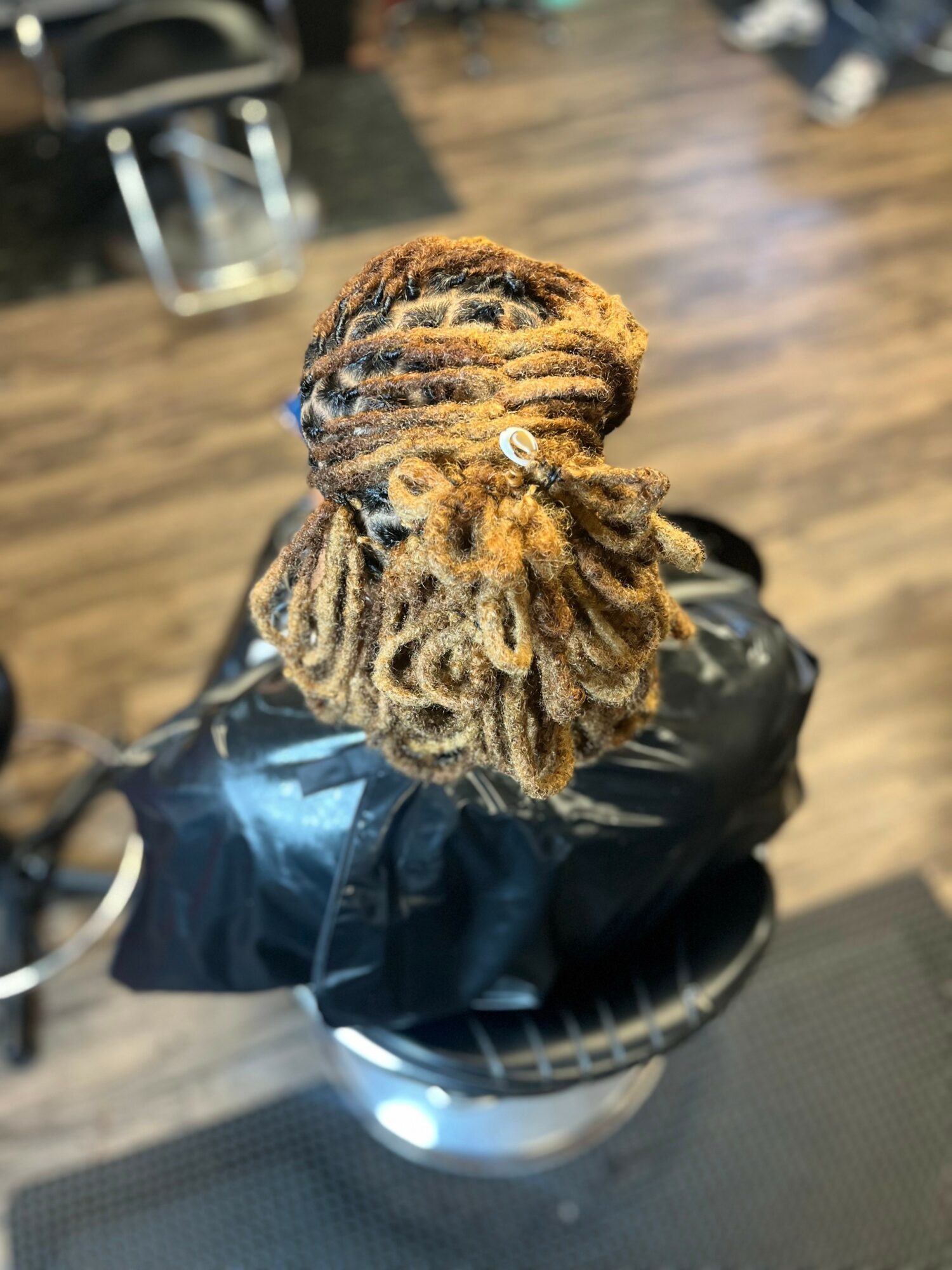 Top view of a person's head with styled dreadlocks, sitting in a salon chair on a wooden floor.