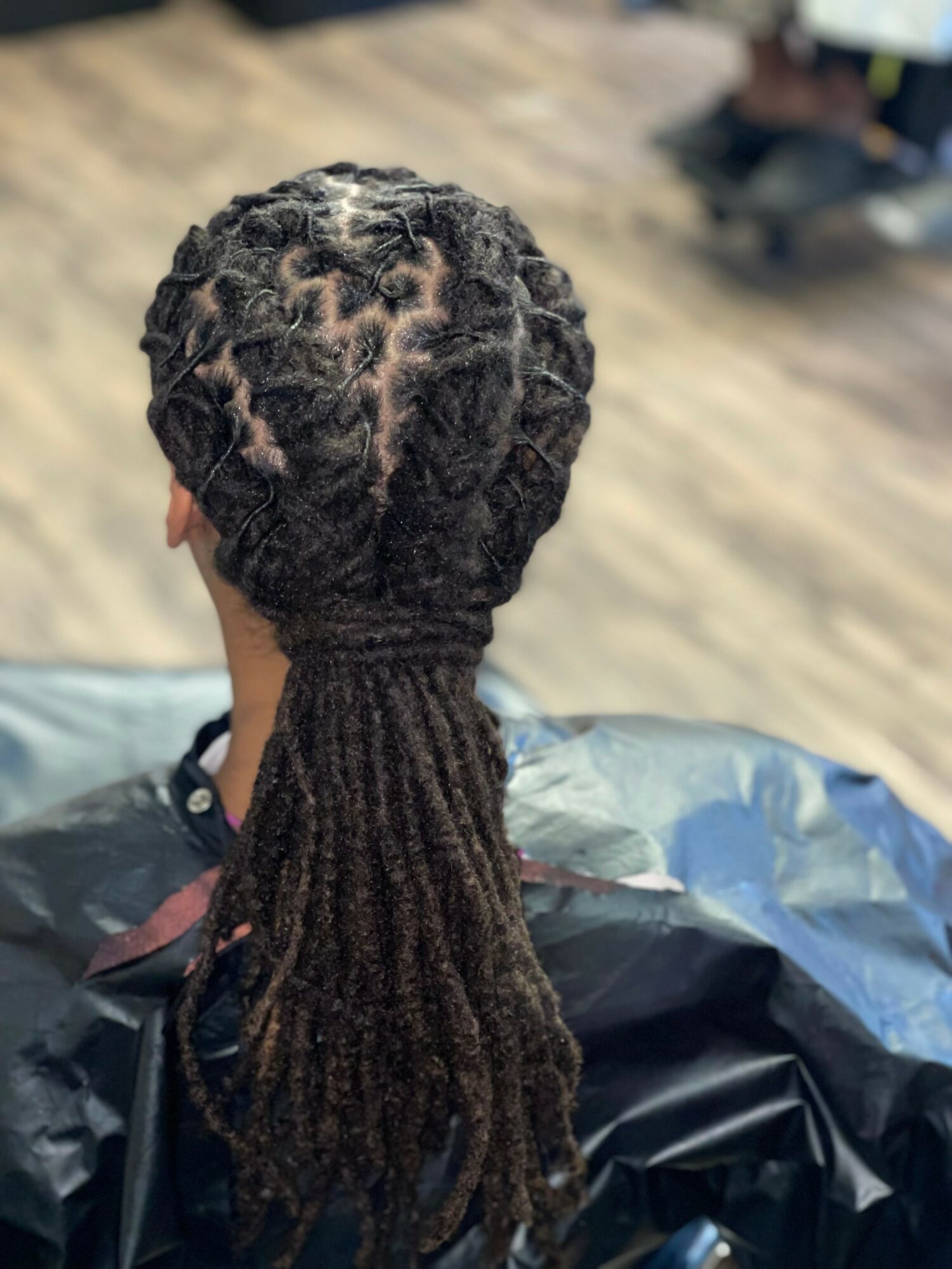 Back view of a person with styled dreadlocks, sitting in a chair, in a room with wooden flooring.