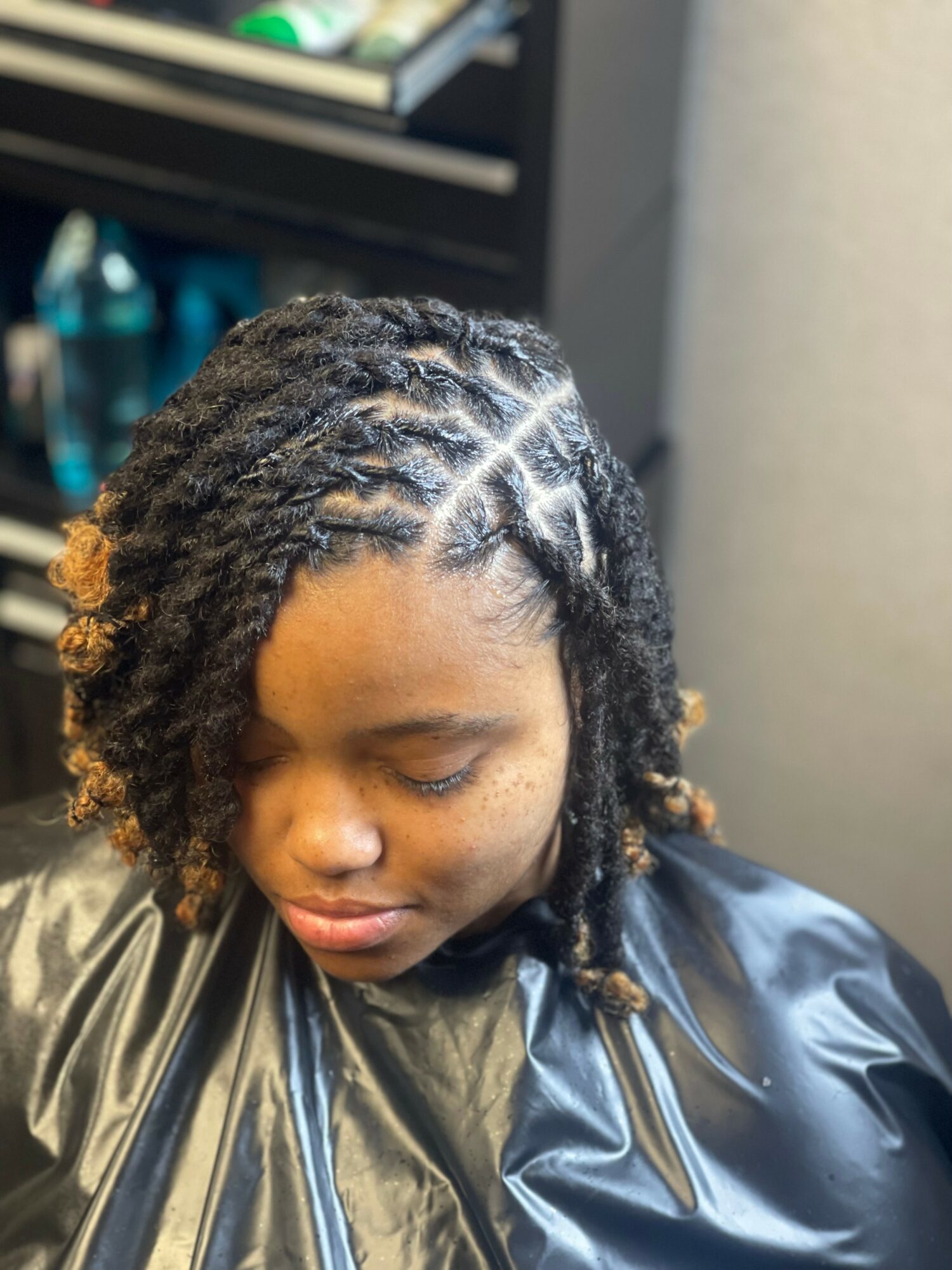 Young woman with styled dreadlocks, wearing a black cape, looking down, in a salon setting.