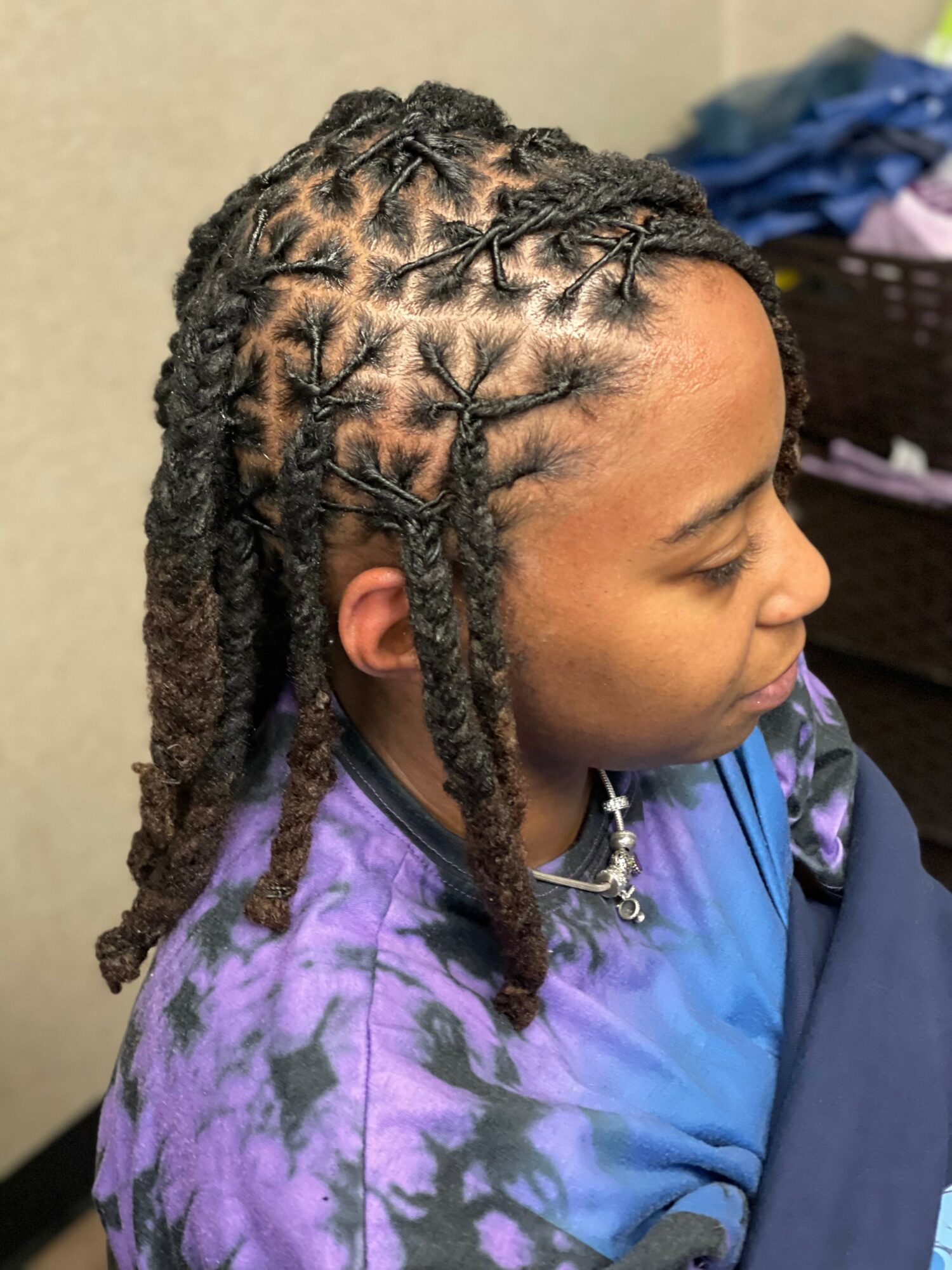 Side view of person with styled dreadlocks and a purple tie-dye shirt, sitting indoors.