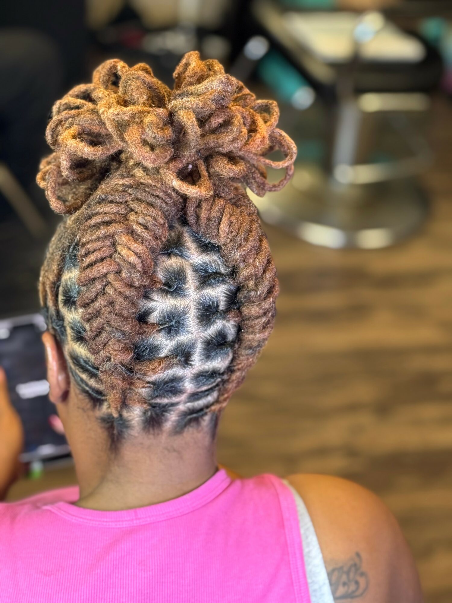 Back of a person's head with styled dreadlocks in a bun, intricate scalp braiding visible, wearing a pink top.