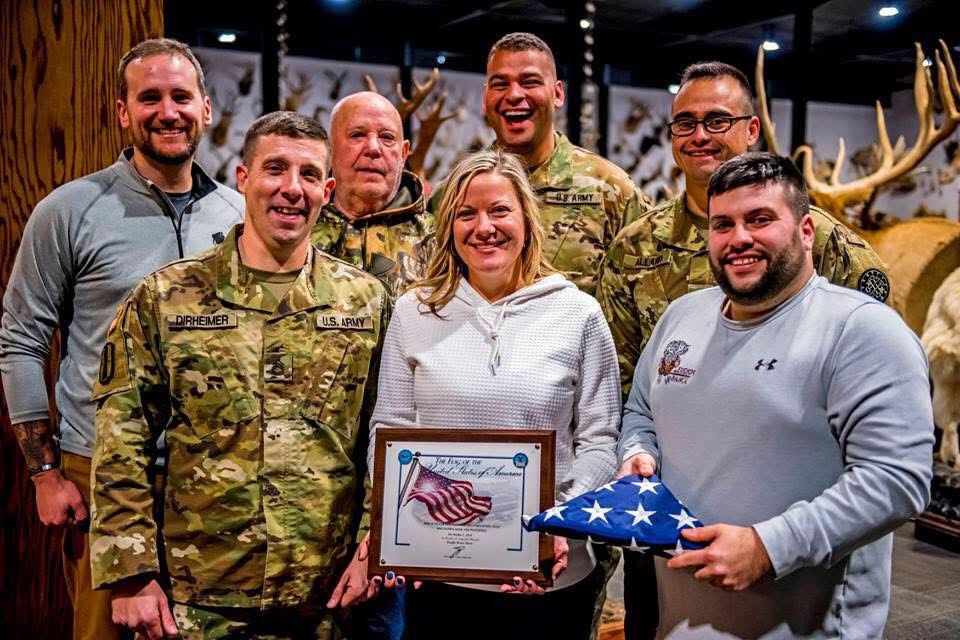 Group of seven people, including military personnel and civilians, smiling indoors with deer antlers in background.