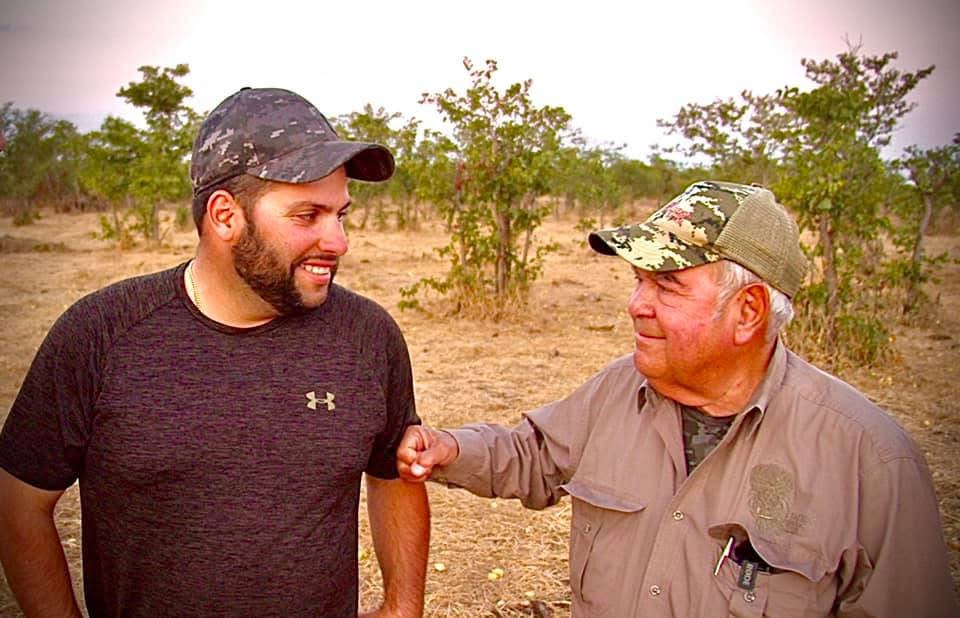 Two men smiling and shaking hands outdoors in a dry, open landscape with trees in the background.