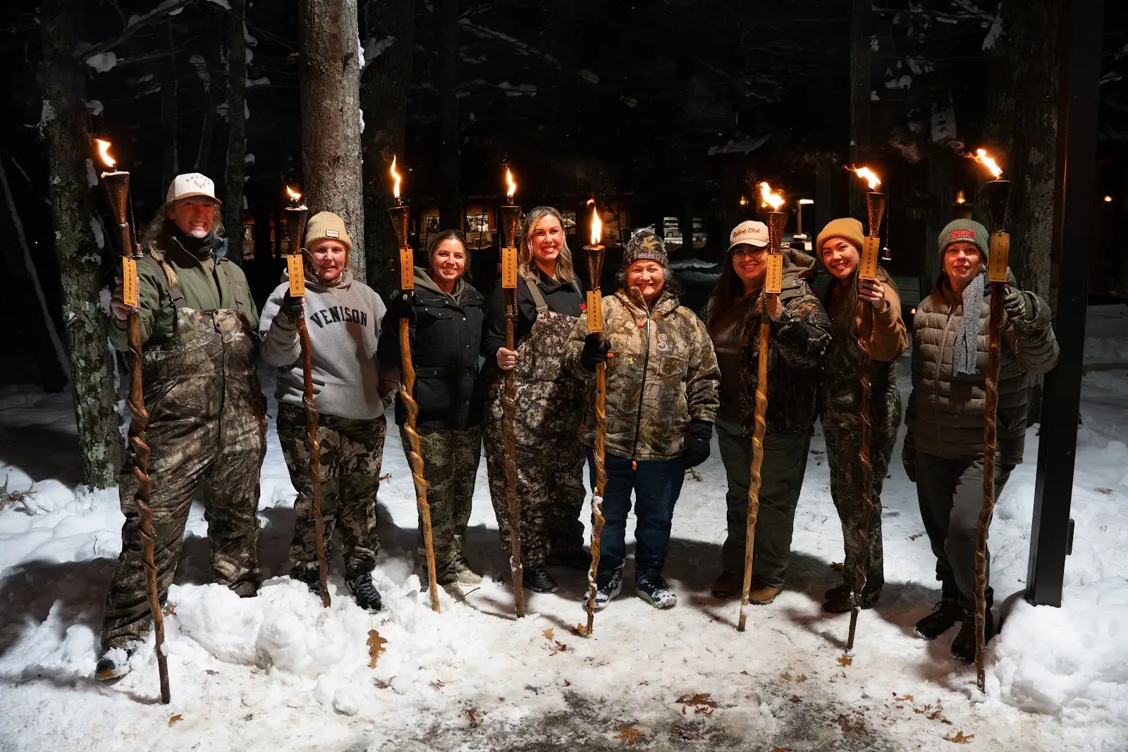 Group of nine people standing outdoors in snow, holding torches, dressed in winter and camouflage clothing, at night.