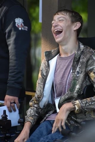 Young person in a wheelchair laughing outdoors with a person standing nearby holding a paper and camera.