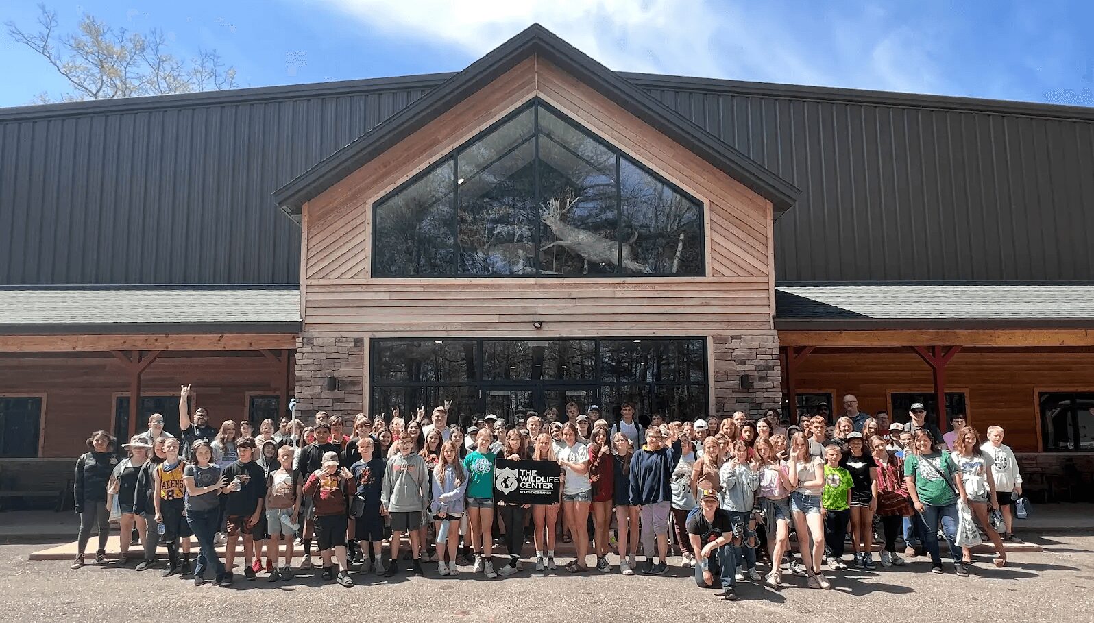 Large group of people gathered outside a building with a glass front and wooden exterior under a blue sky.