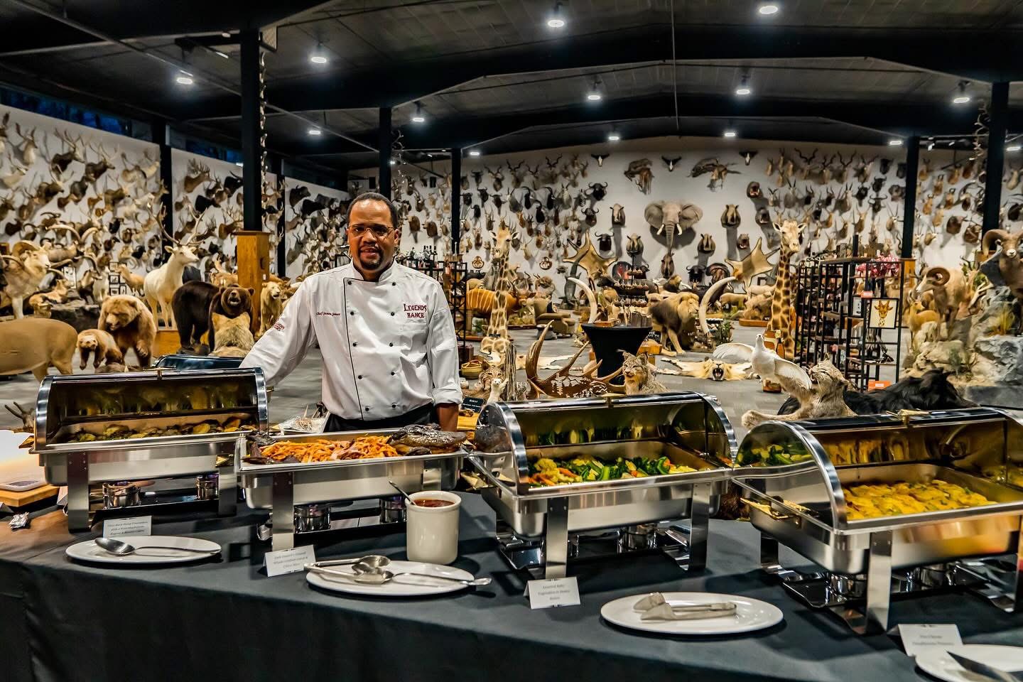 Chef standing behind a buffet with various food trays in a decorated room with animal displays on the wall.