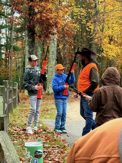 Three people outdoors in a wooded area with autumn leaves, two children holding orange and black objects, an adult in an orange vest, and a person in a brown jacket.
