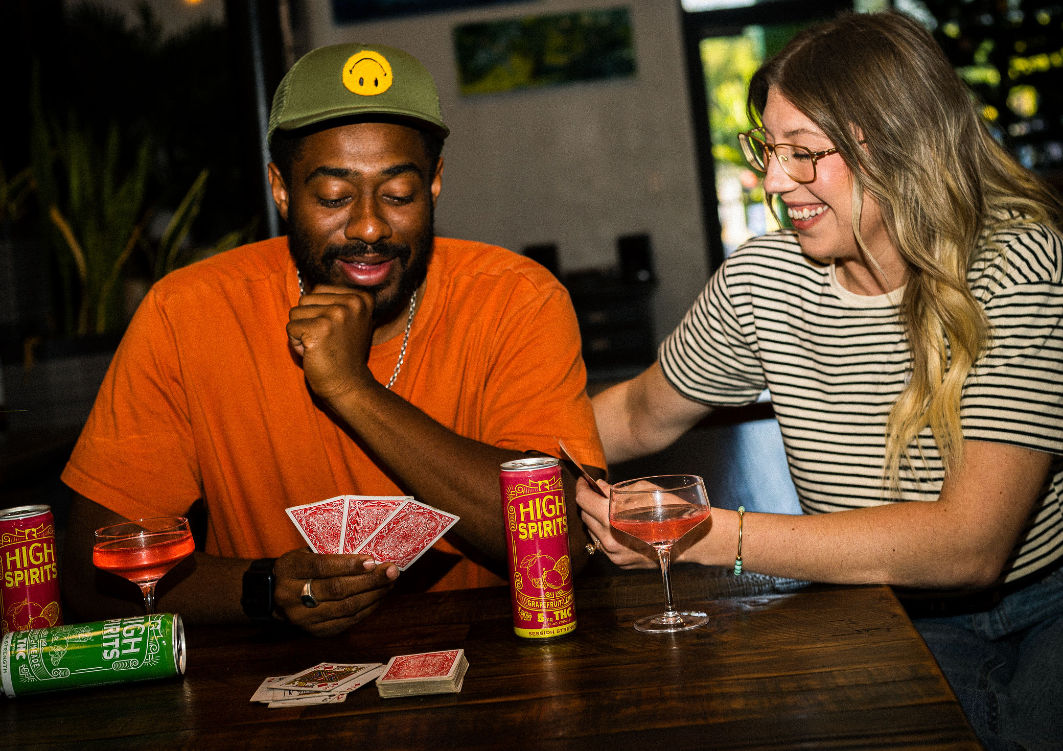 Two people playing cards at a table, smiling, with drinks and snacks, in a casual setting.