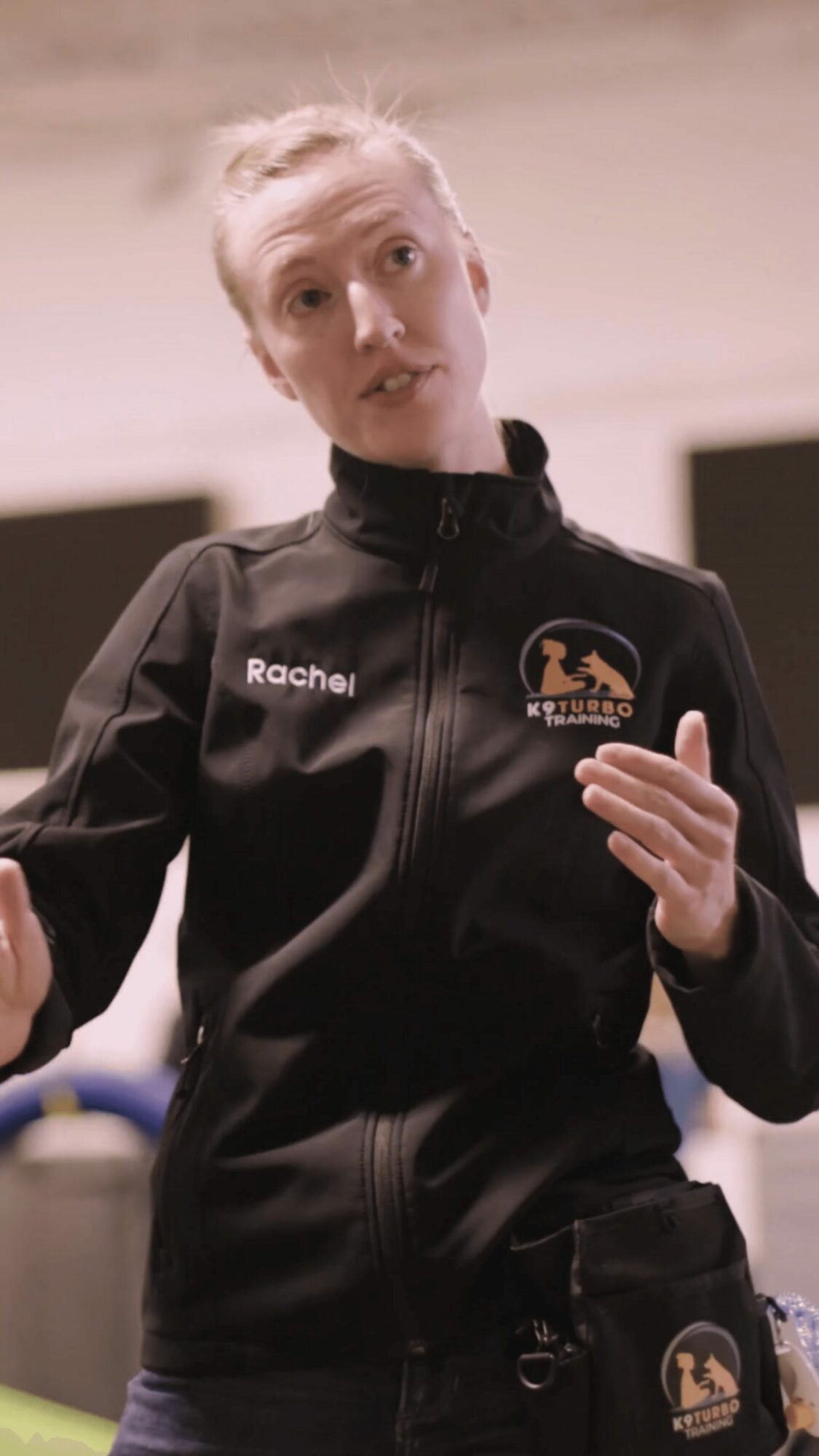 Young woman with short blonde hair wearing a black jacket with logos, speaking and gesturing with her hands.
