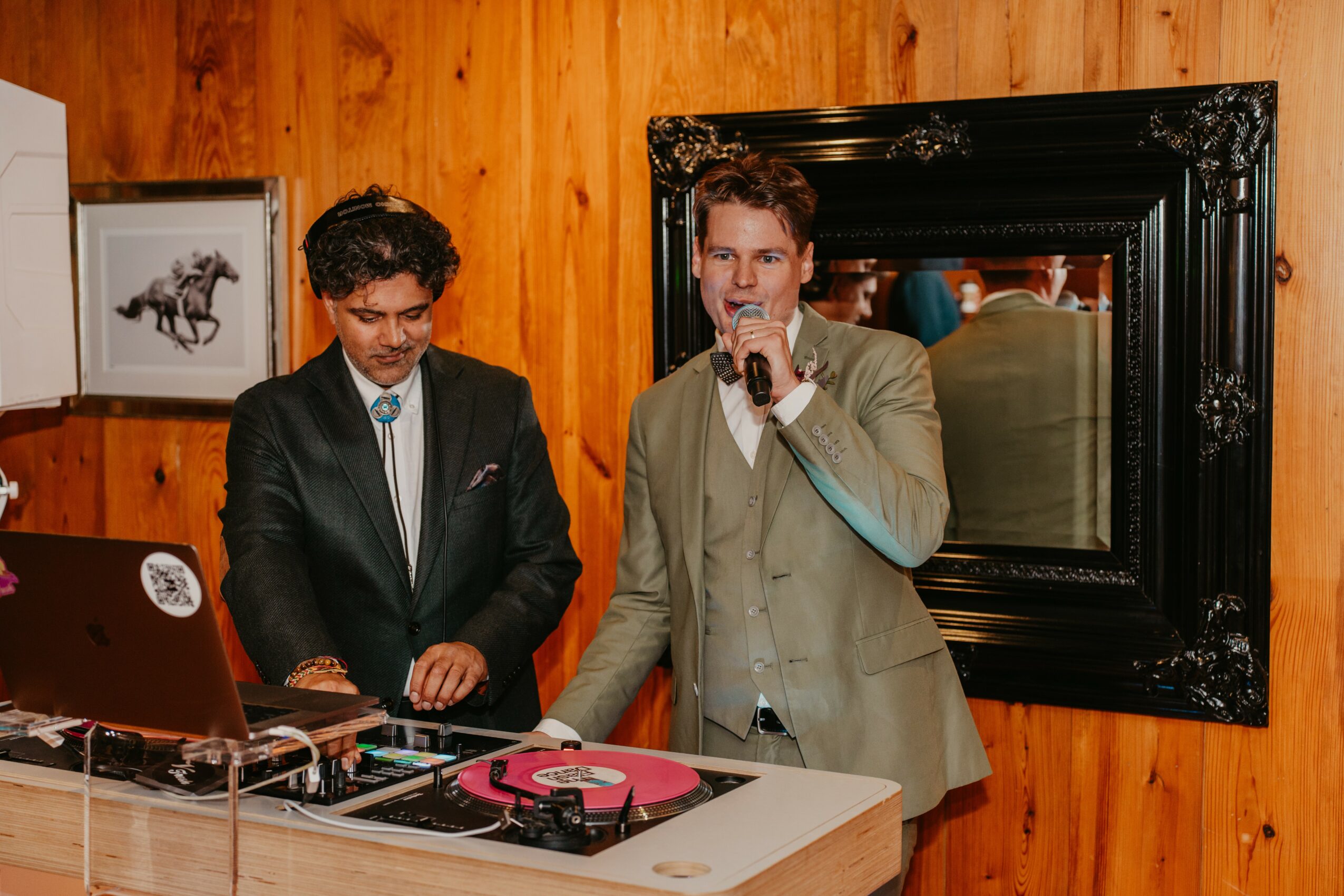 Two men in suits stand at a table with DJ equipment, one speaking into a microphone, wooden wall background.