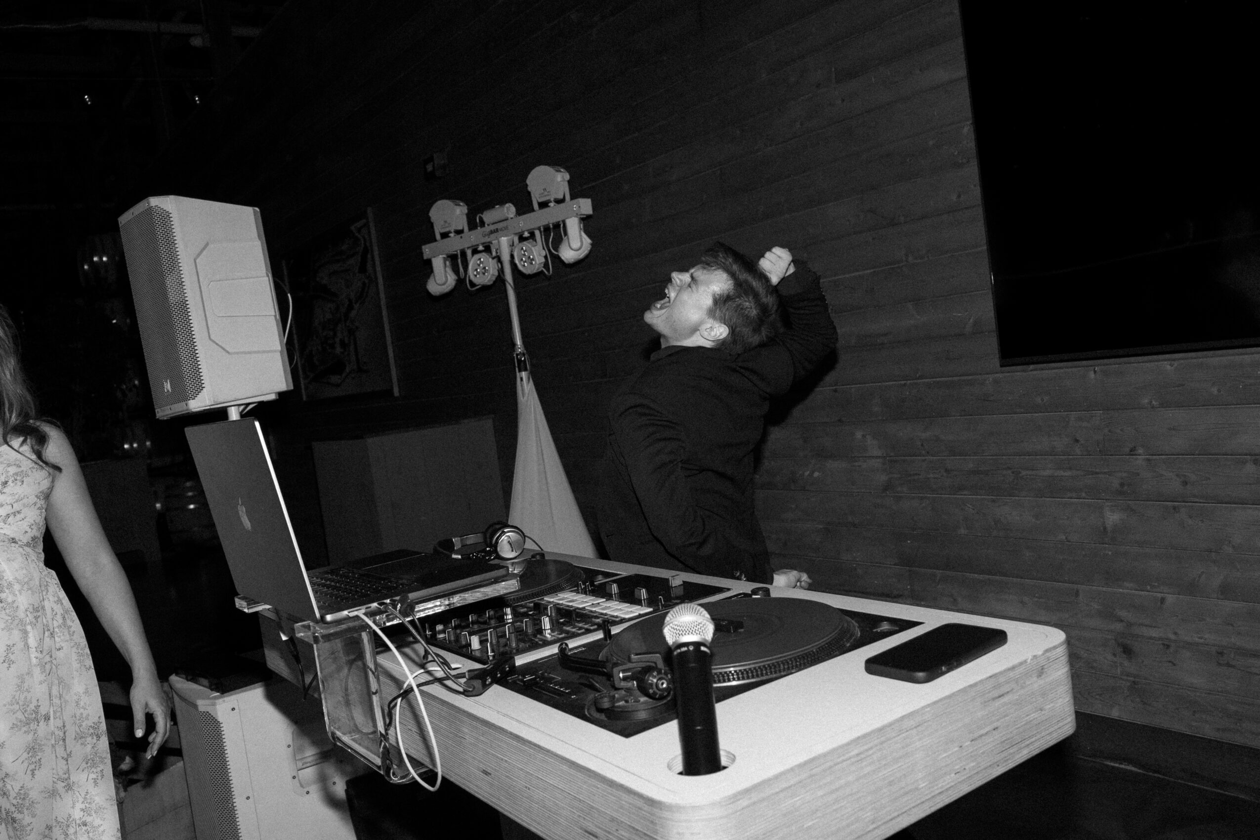 Person with hand on head standing behind DJ equipment in a dark room with wooden wall.