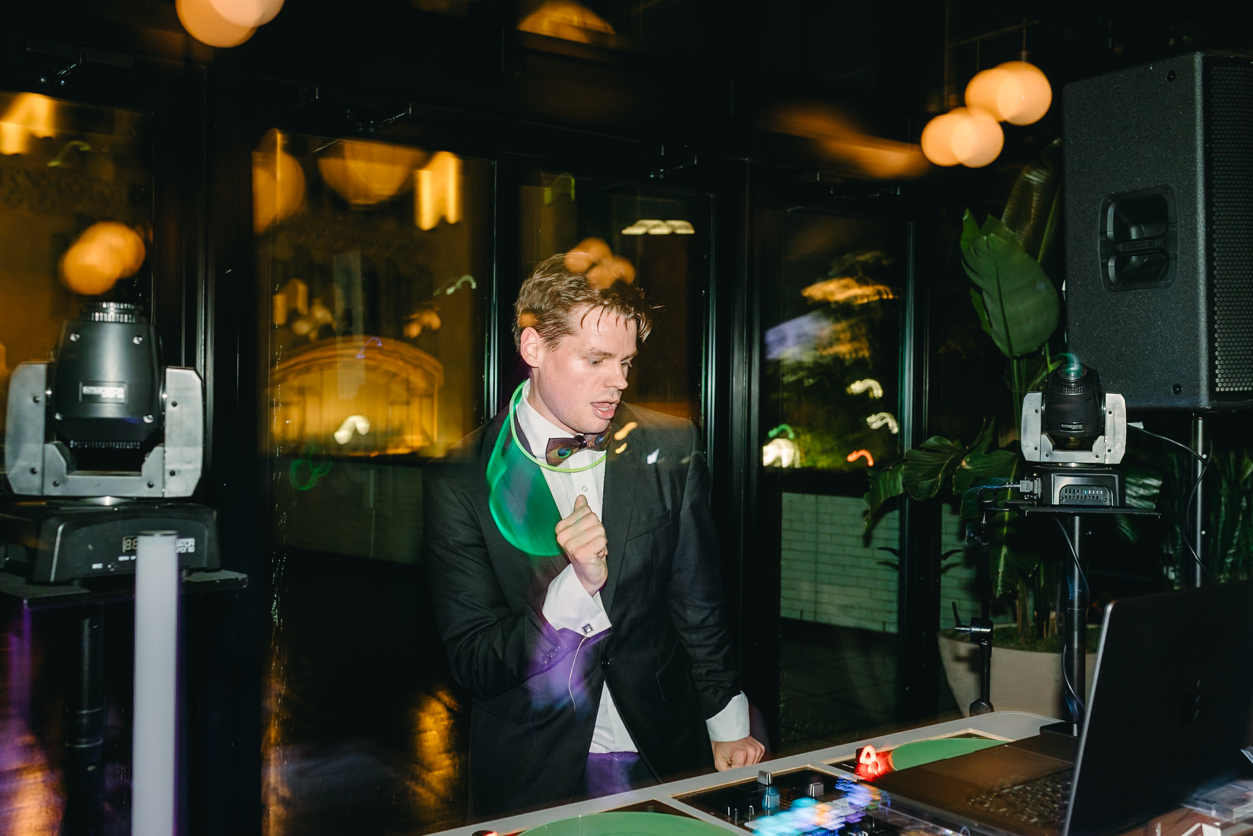 Young man in a tuxedo with a green scarf holding a microphone, standing near a laptop and speakers in a dimly lit room.