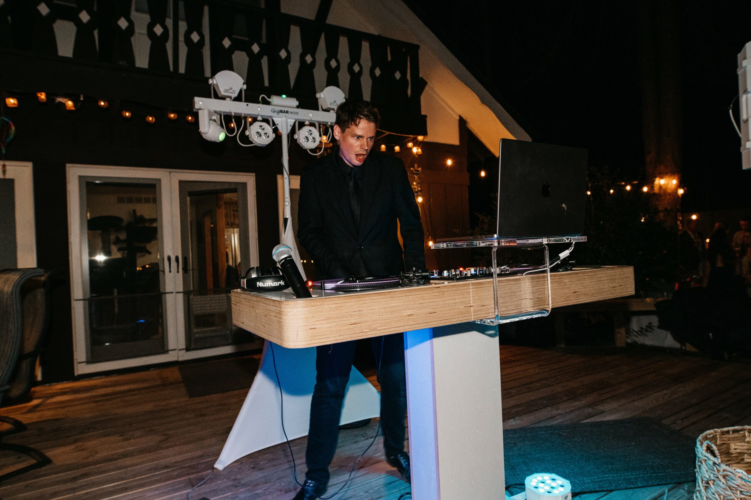Person standing behind a DJ setup with equipment and laptop, indoors with warm lighting and decor.
