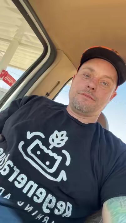 Man wearing a black cap and T-shirt sitting inside a vehicle, looking at the camera.