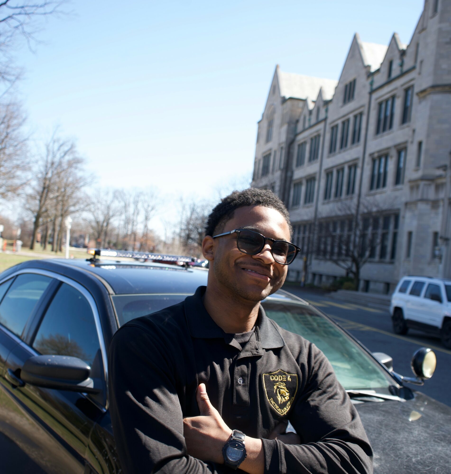 Young man wearing sunglasses and a black jacket with a badge, standing outdoors near a car, smiling.