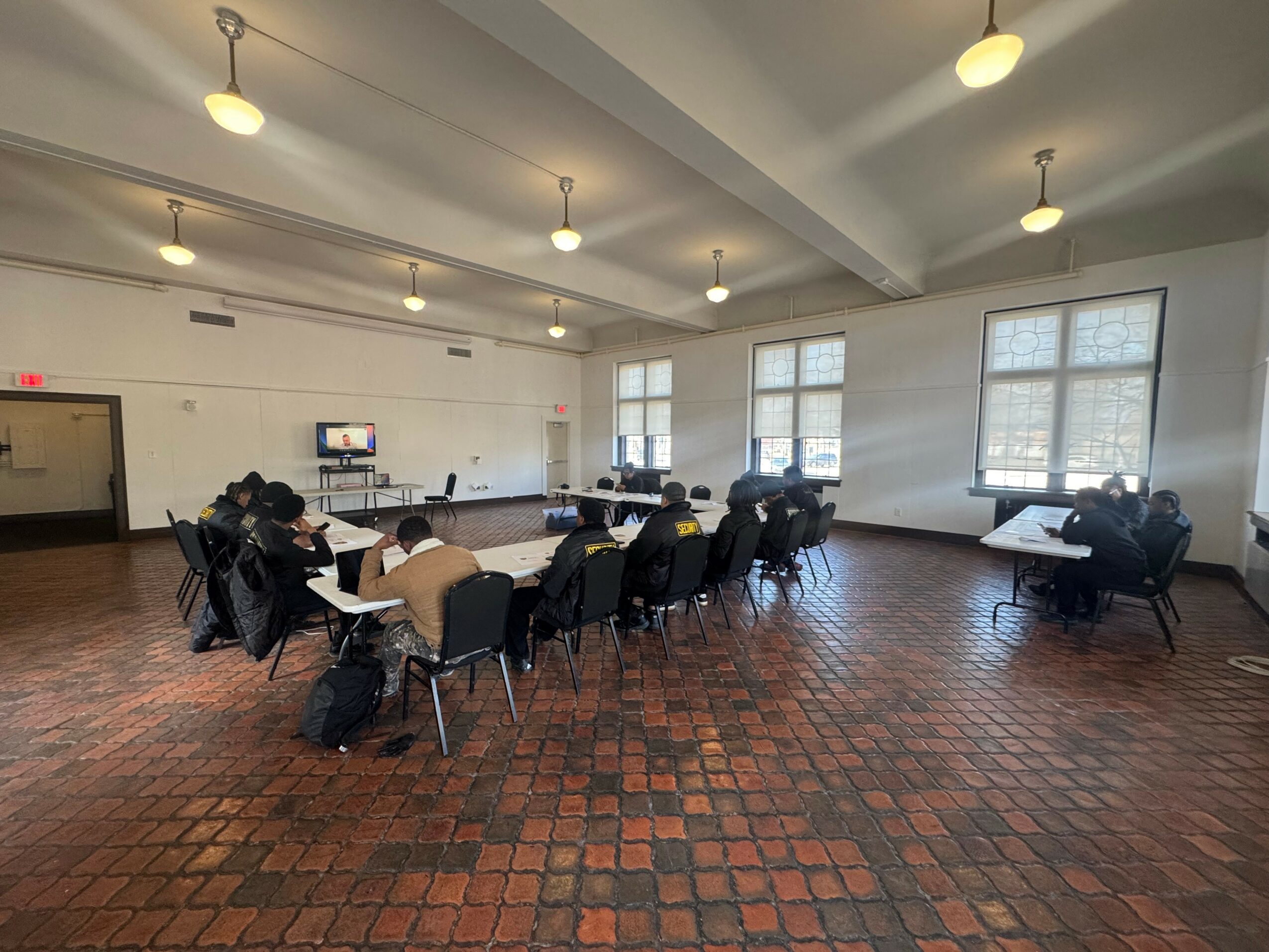Large room with tables and chairs, windows with blinds, and ceiling lights, possibly a classroom or meeting space.