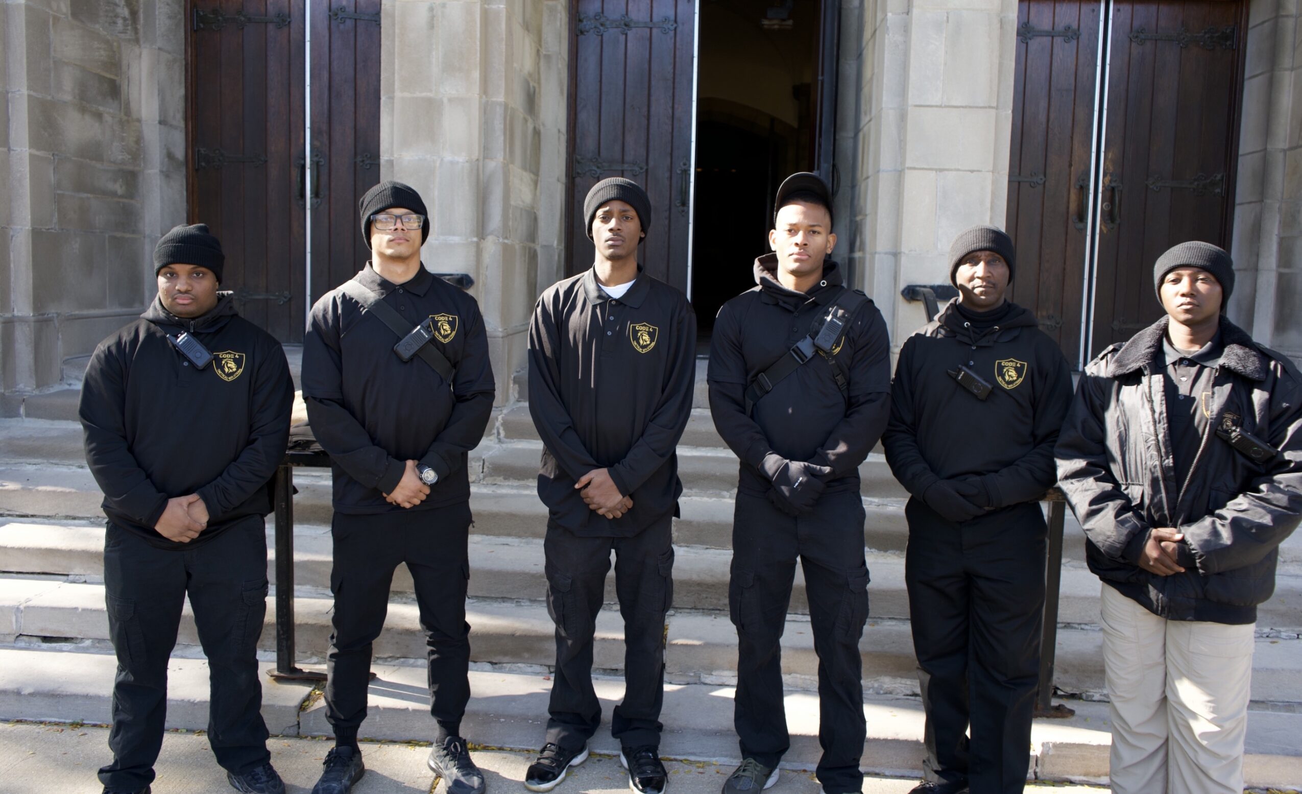 Six people stand in front of a building, wearing dark clothing and hats, some with hands in pockets.
