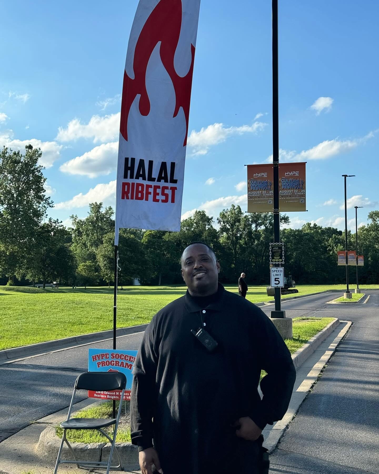 Man standing outdoors near a flag with flames and text, with trees and a parking lot in the background.