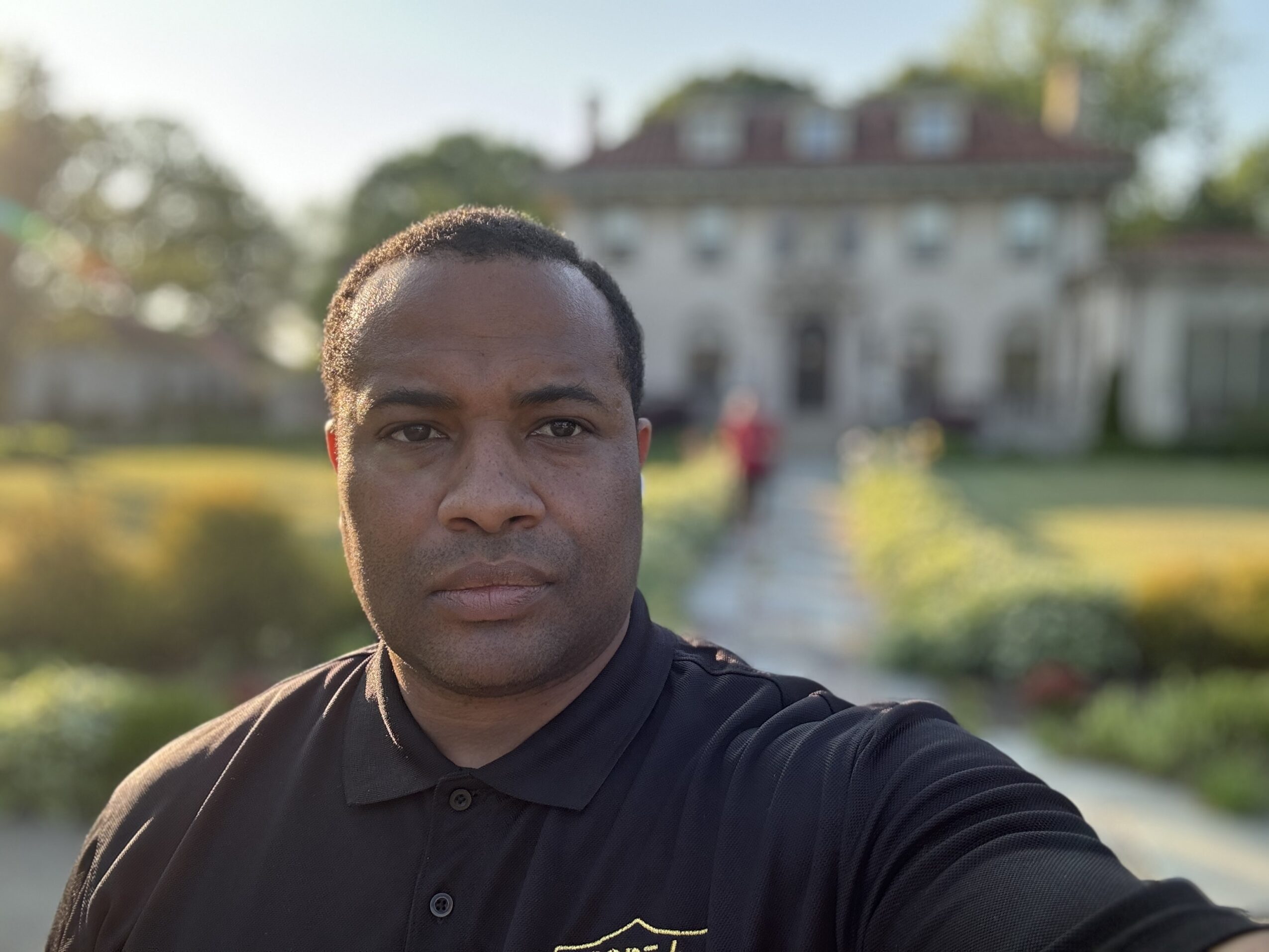Man taking selfie outdoors with a building and garden in background.