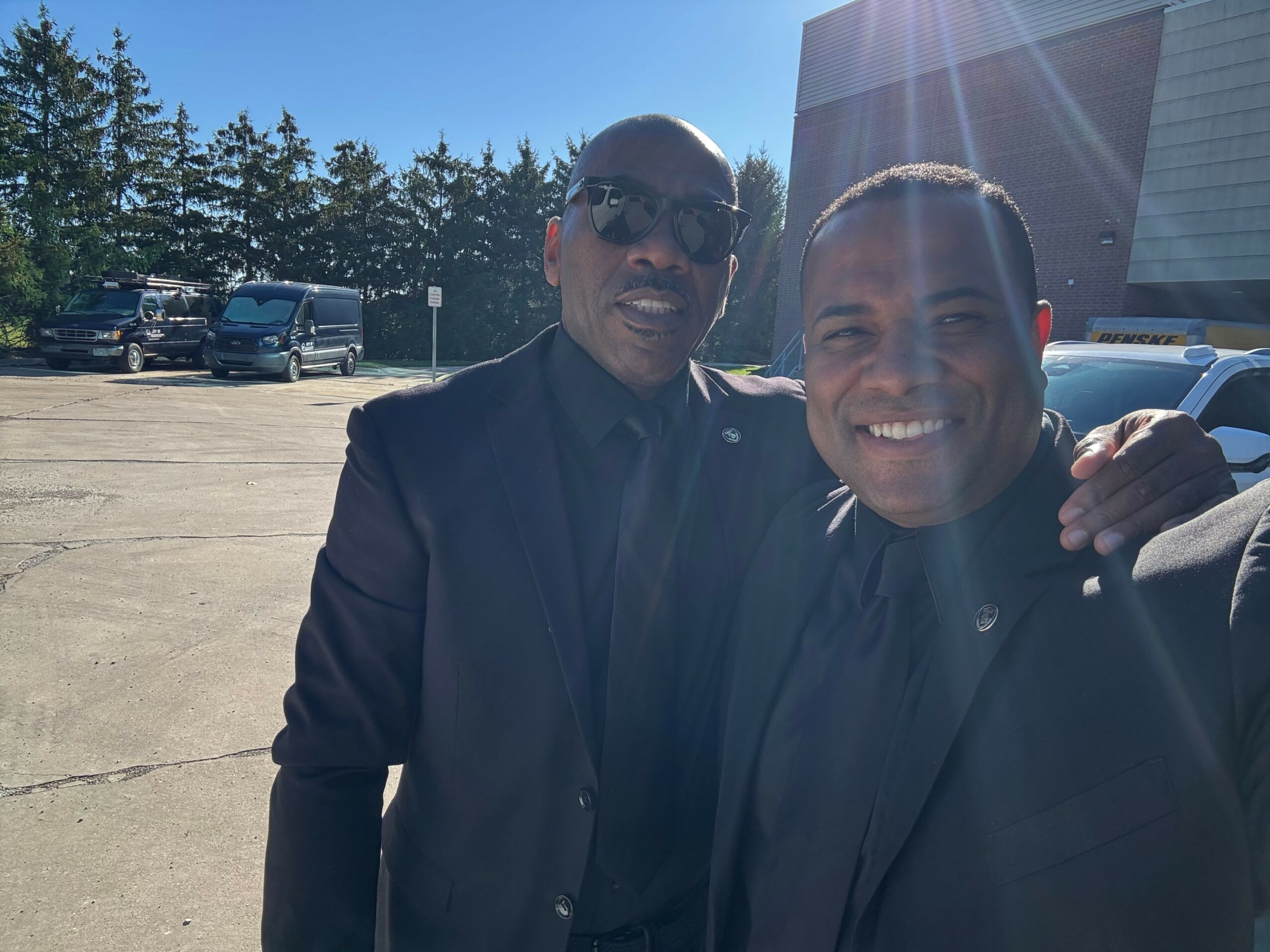 Two men in suits smiling outdoors with parked cars and trees in background.