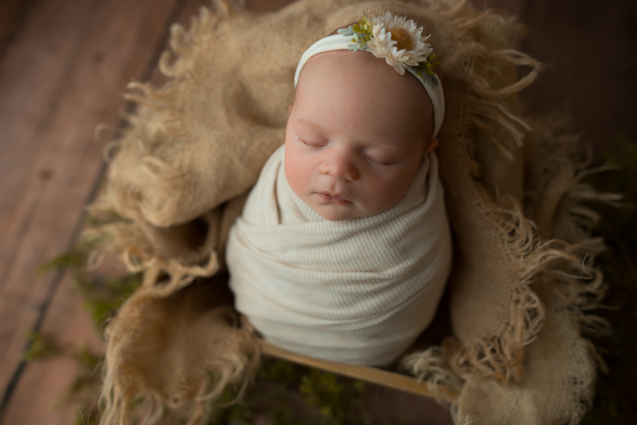 Baby with a daisy headband wrapped in a white blanket, lying on a soft blanket, eyes closed.