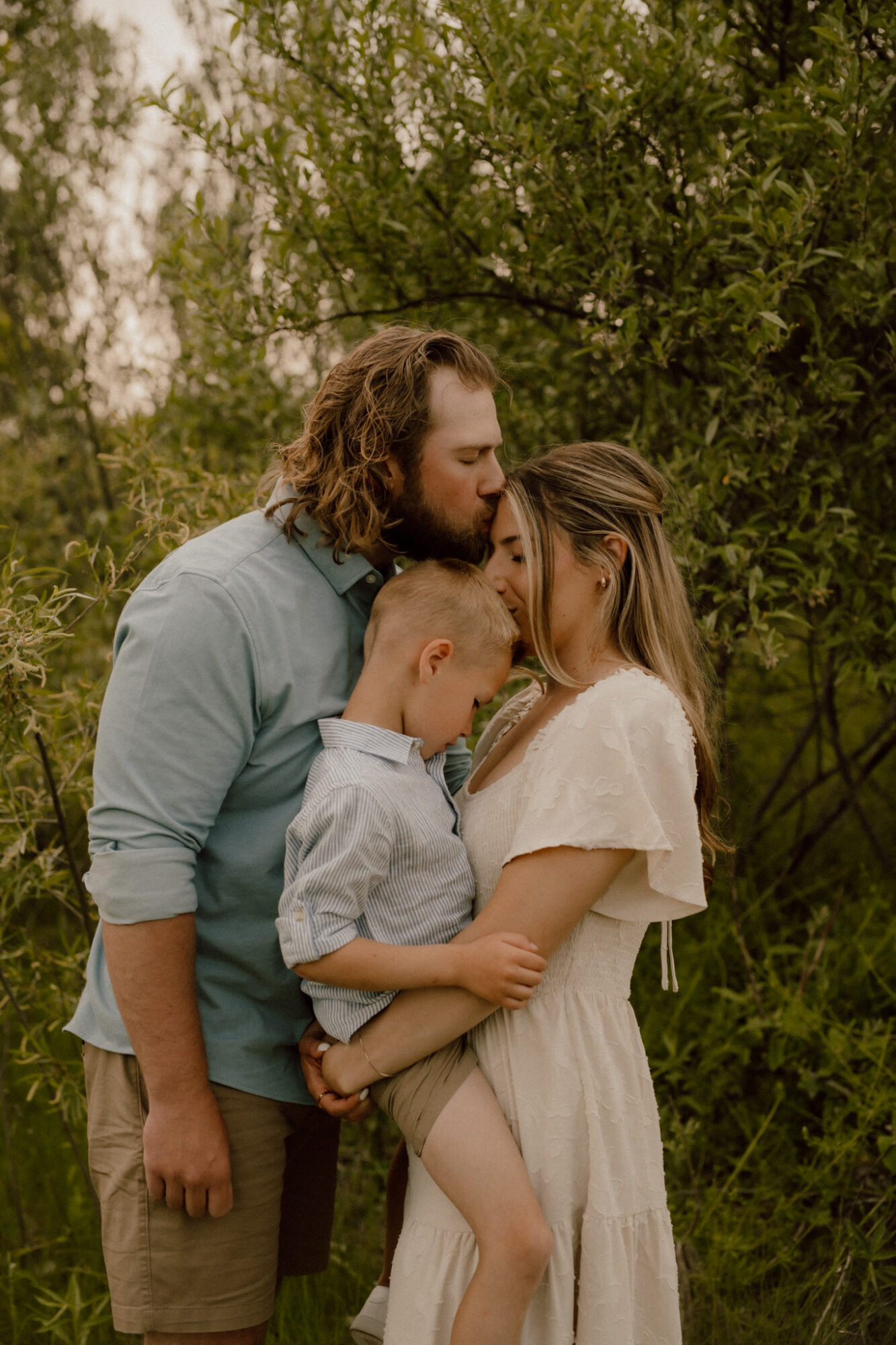 A family of three stands outdoors surrounded by green trees, embracing closely and sharing a tender moment.