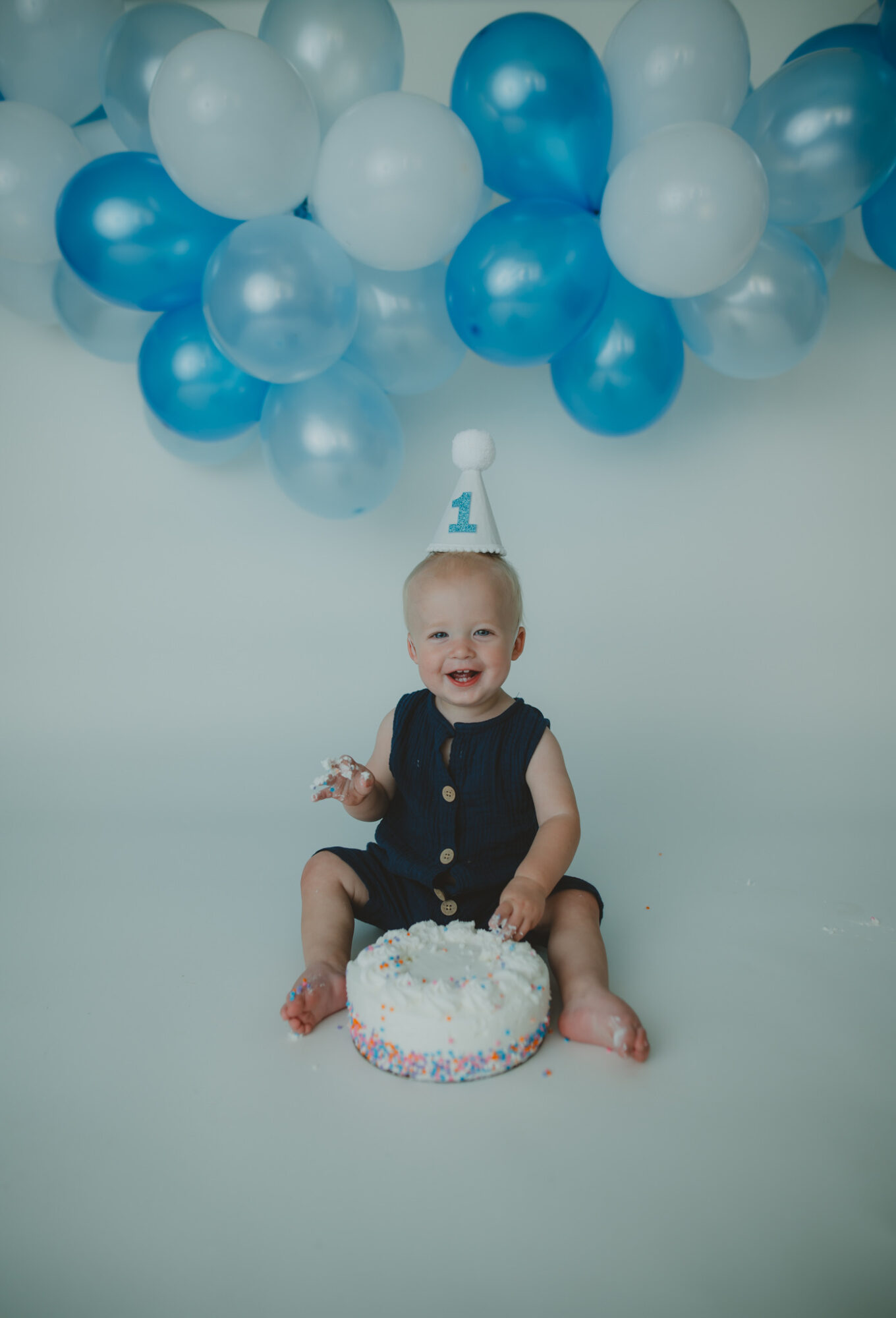 Smiling baby sitting on the floor with a cake, surrounded by blue and white balloons, celebrating first birthday.