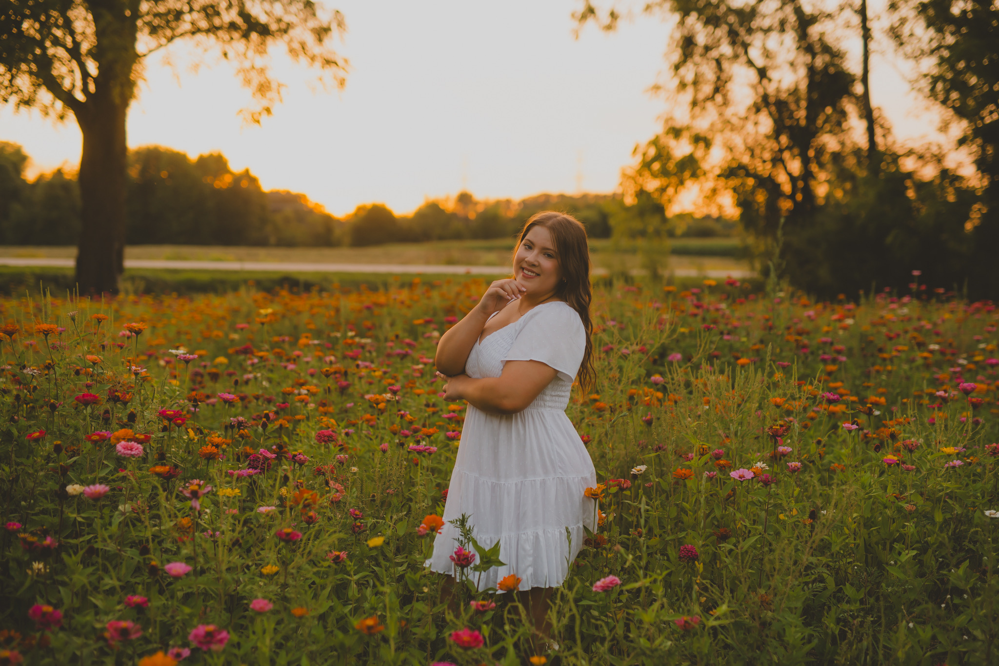 Woman in white dress standing in a flower field during sunset, smiling with arms crossed.