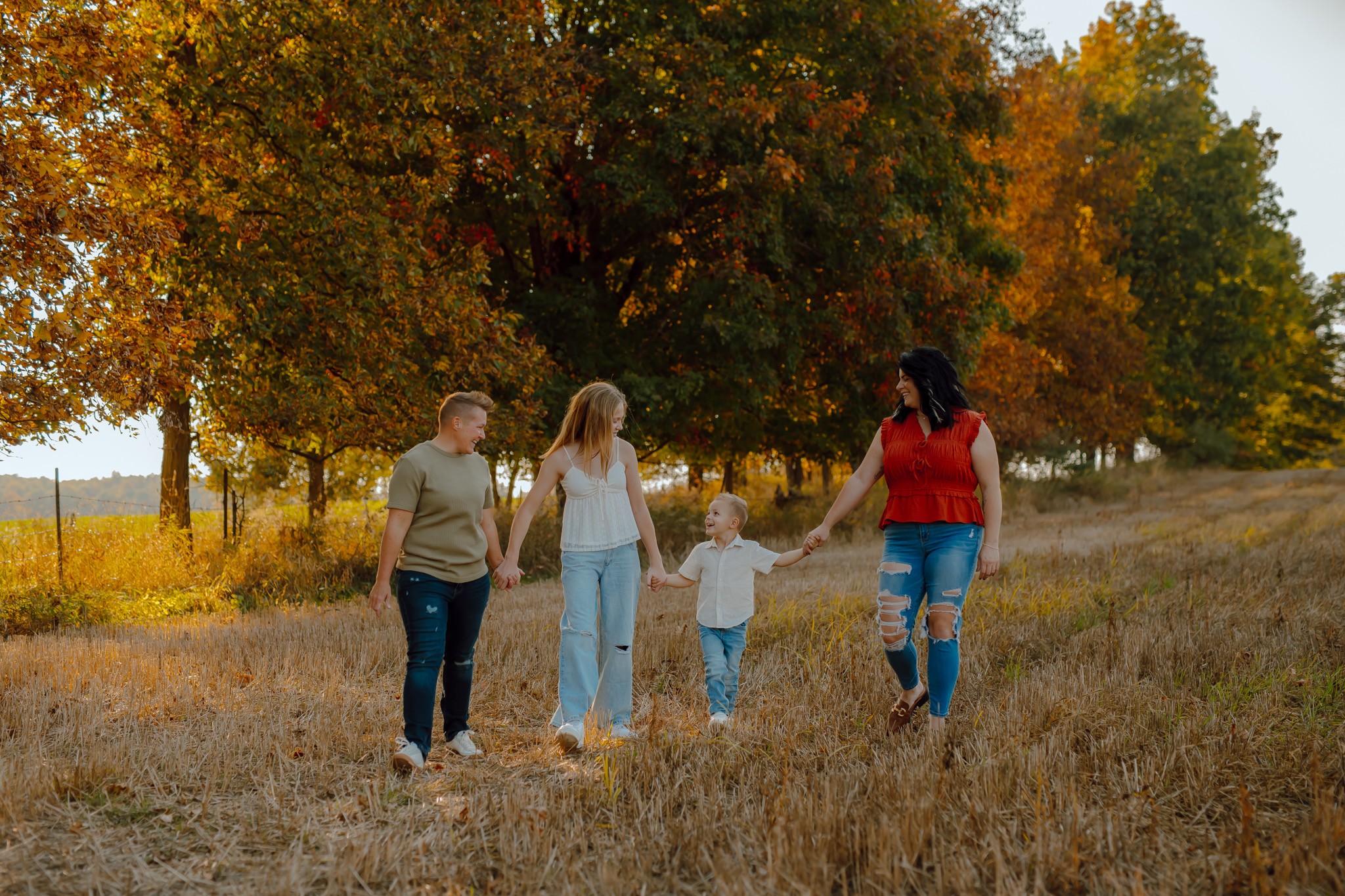 Four people walking through a grassy field with trees in autumn colors in the background.