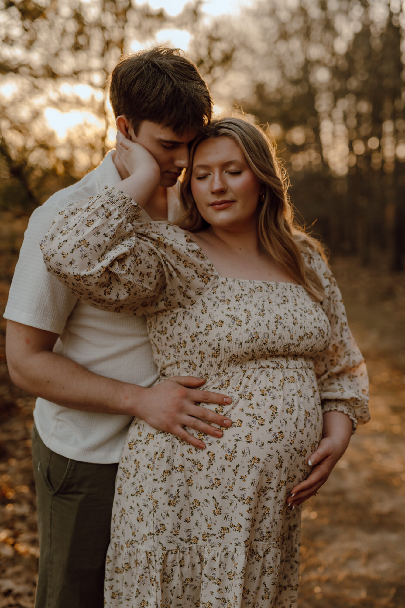 A pregnant woman and a man stand outdoors during sunset, embracing each other gently with trees in the background.