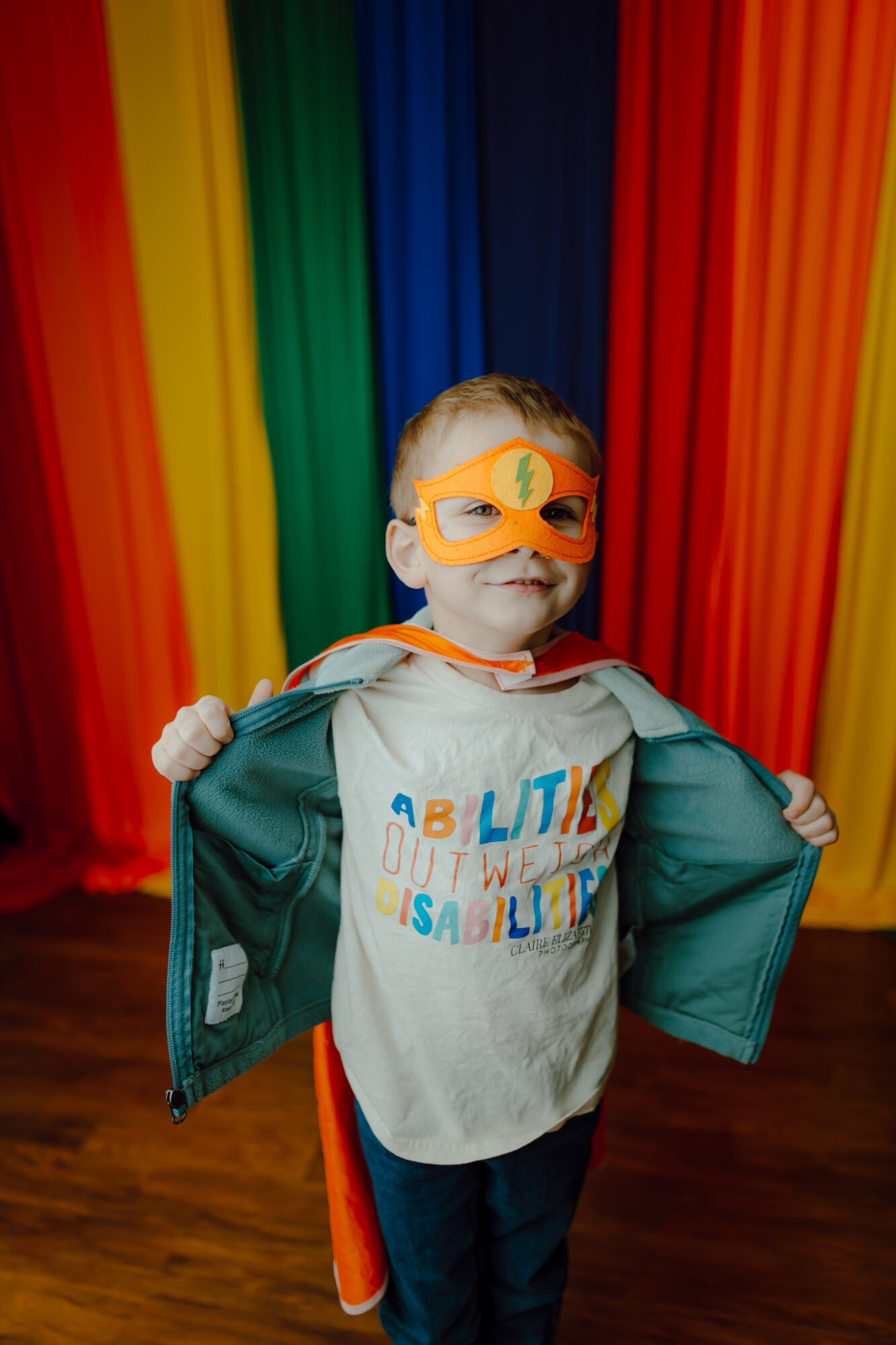 Child wearing orange mask and cape, holding open jacket, standing in front of rainbow-colored curtains.