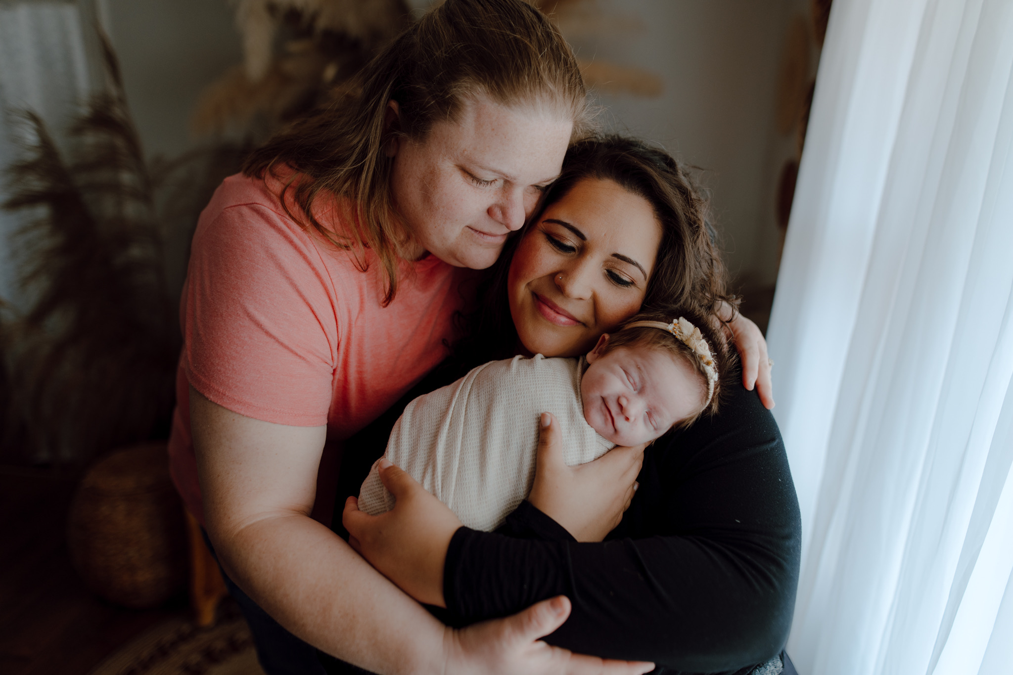 Two women hold a newborn baby near a window with curtains, smiling and embracing each other.