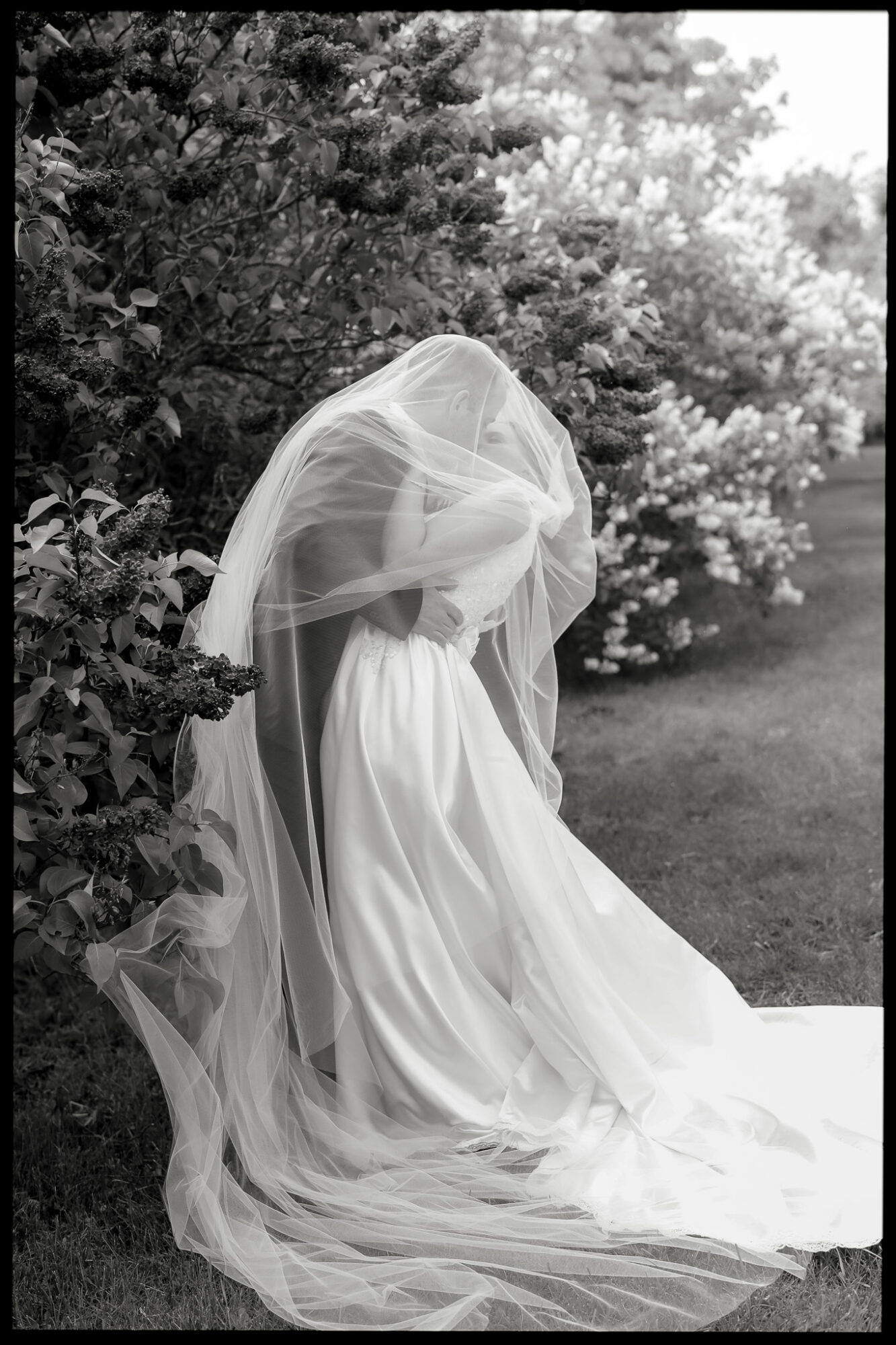 Person in wedding dress and veil sitting outdoors near bushes, with trees and grass in background.
