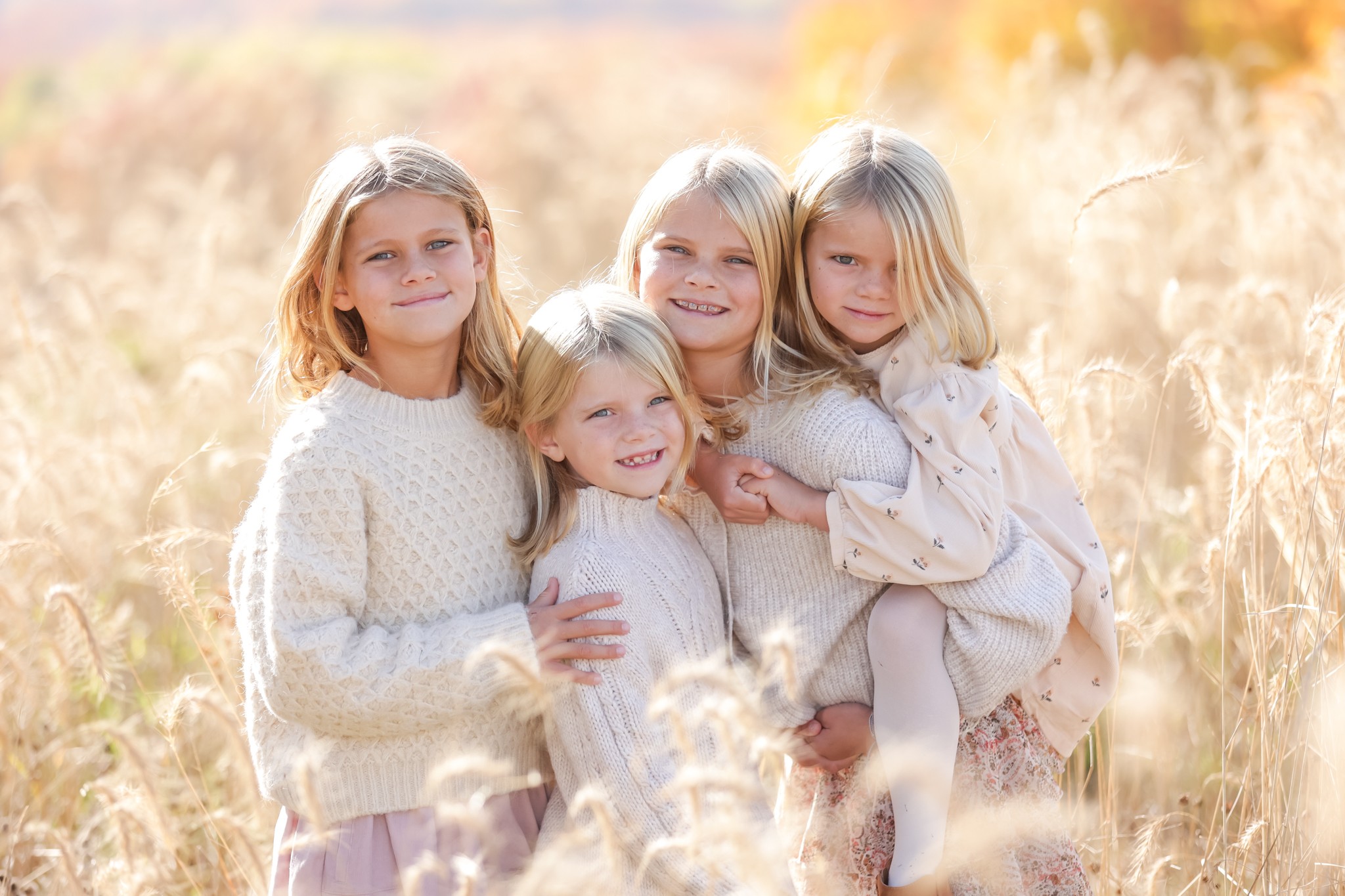 Four young girls with blonde hair smiling in a field of tall grass during daytime.