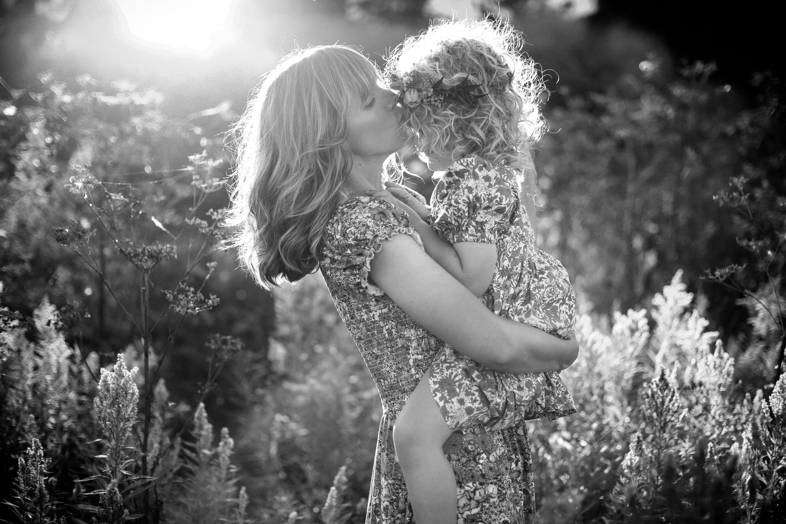 Woman holding a child outdoors with sunlight and plants in the background.