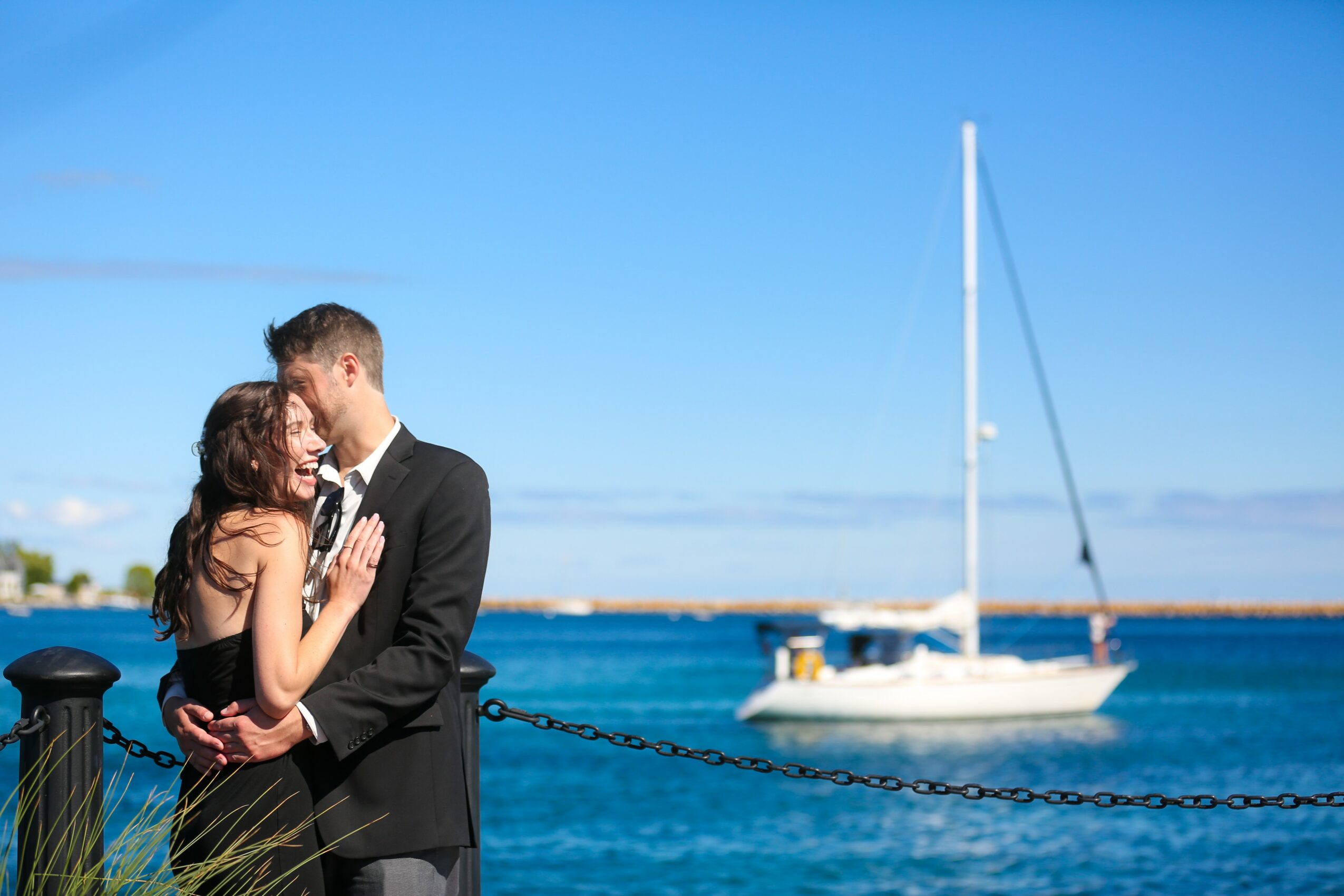 A couple embracing near a body of water with a sailboat in the background under a clear blue sky.